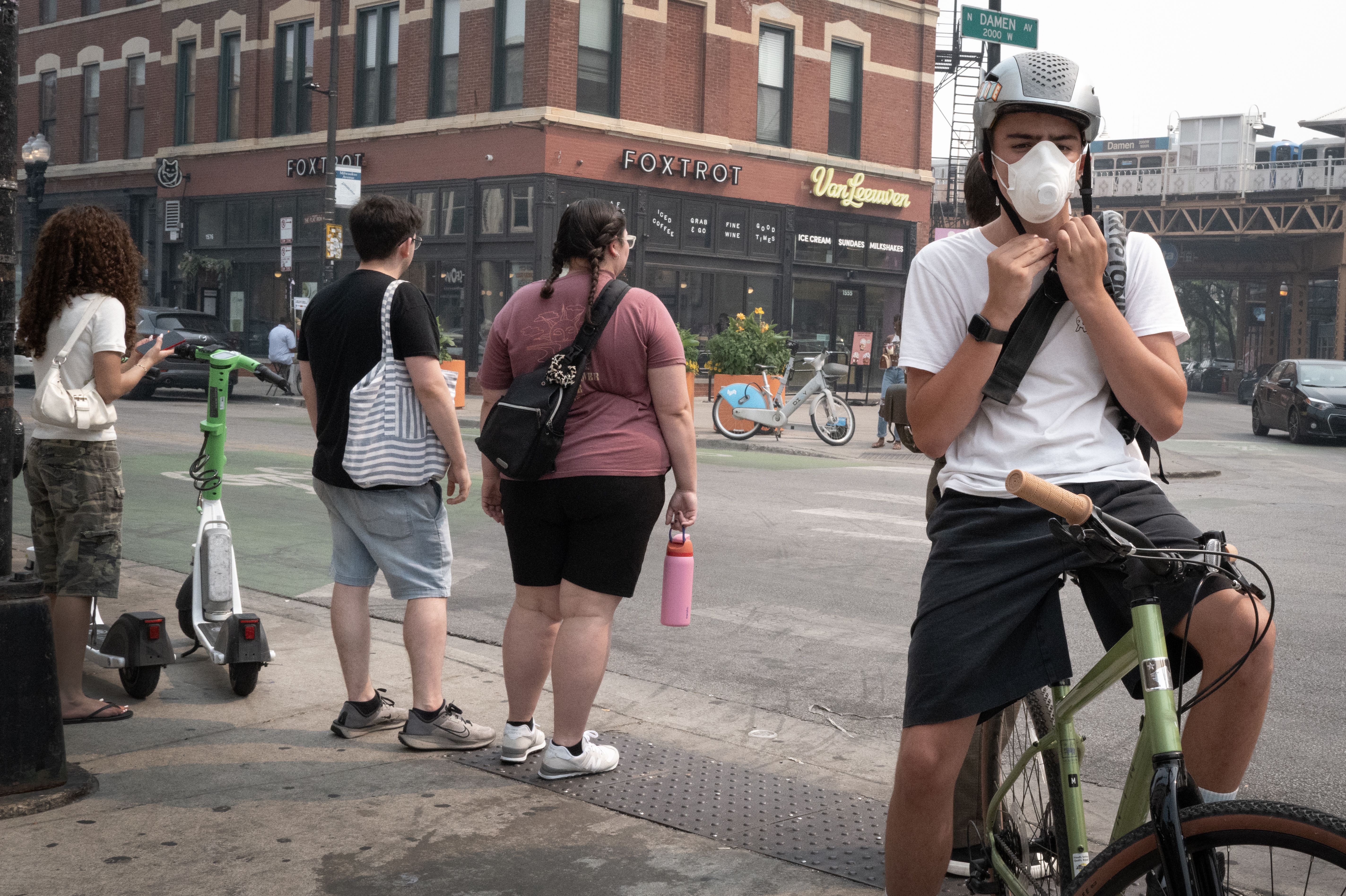 Pedestrians on a city sidewalk by a brick building with a Foxtrot sign; a masked cyclist on a green bike is at the right, others carry bags and a pink water bottle, with a green scooter nearby.