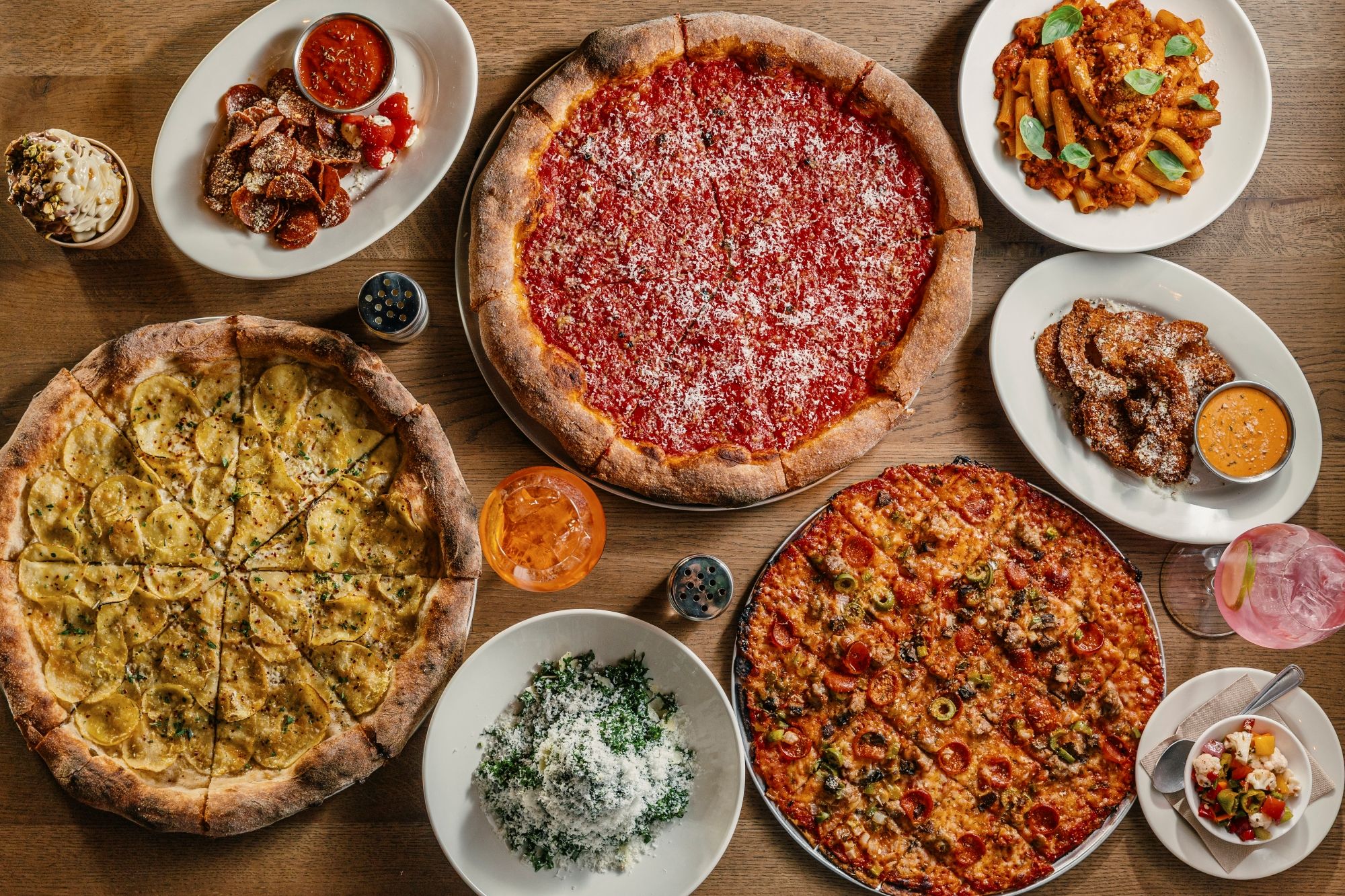 Overhead view of a wooden table jam-packed with pizzas and sides: center is pepperoni pizza, flanked by a white potato-topped pizza and other pies, plus pasta, fried bites with dip, and a dessert cup.