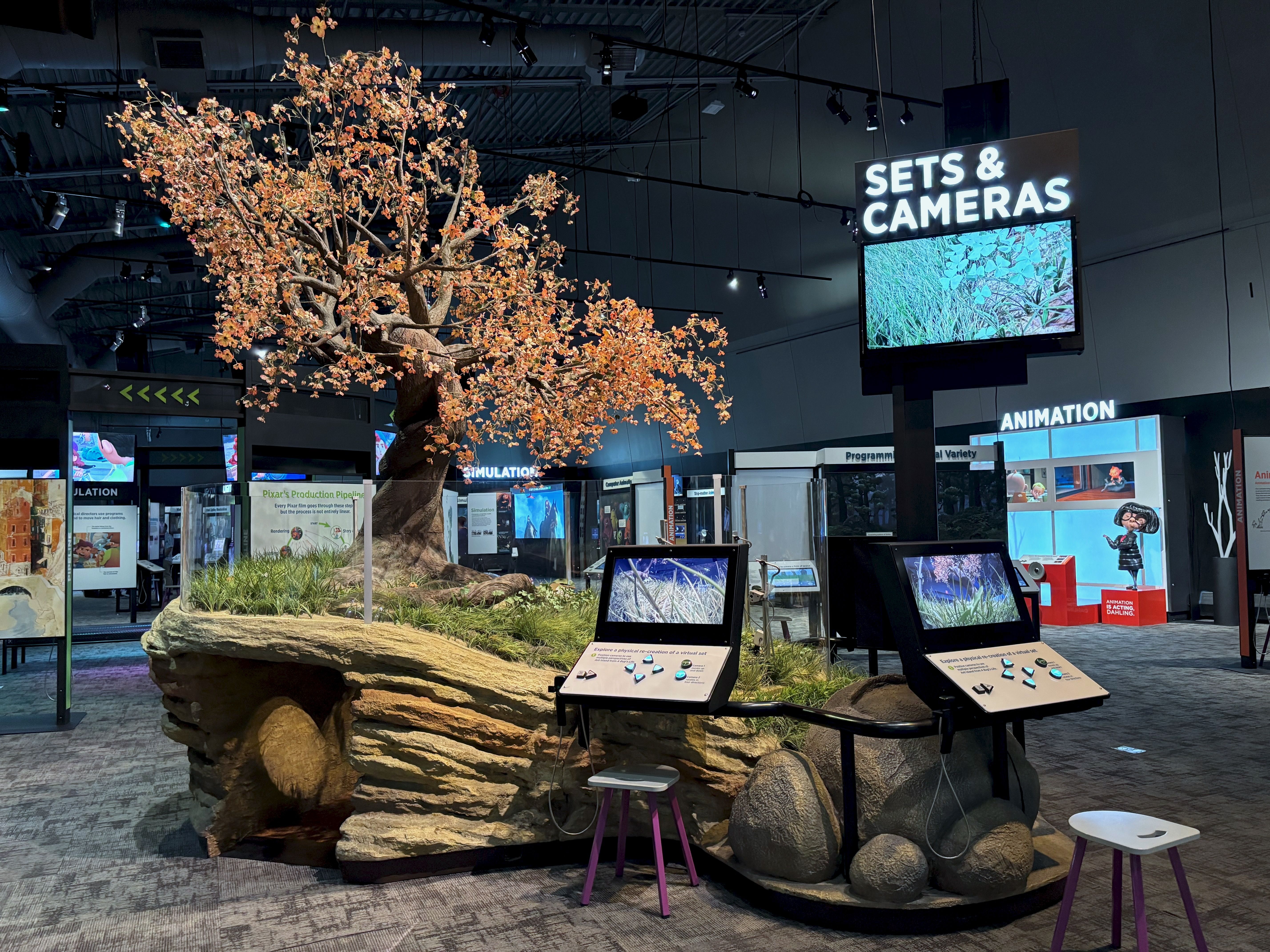 A recreation of Ant Island from "A Bug's Life," featuring an artificial tree with orange leaves, a faux rock cave, interactive screens, and a sign reading "SETS & CAMERAS" with an animation section visible in the background.