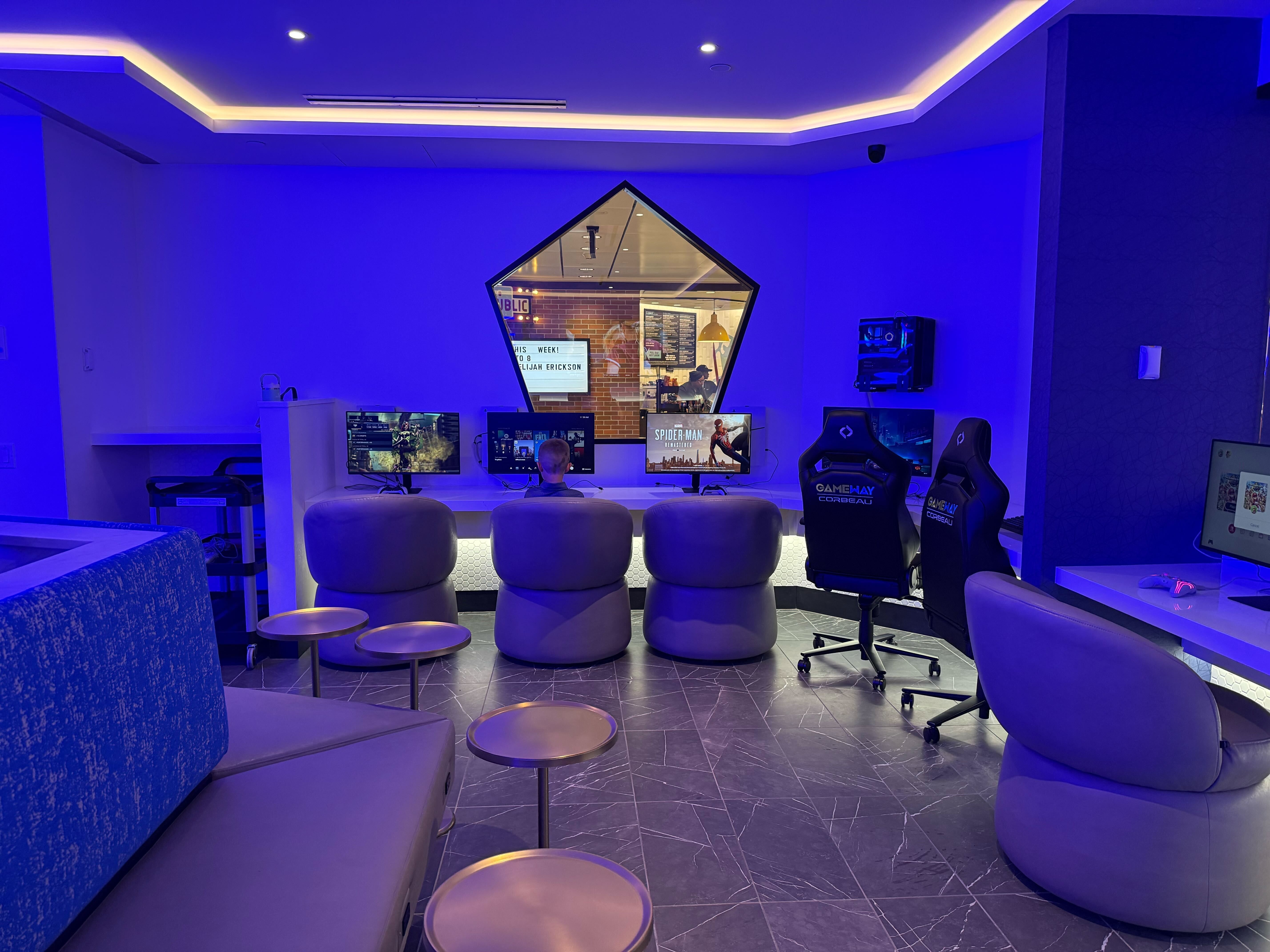 Blue-lit esports lounge with a curved sofa, row of desks and monitors, gaming chairs, and small round stools. A hexagonal window shows a brick-walled room with people at computers.