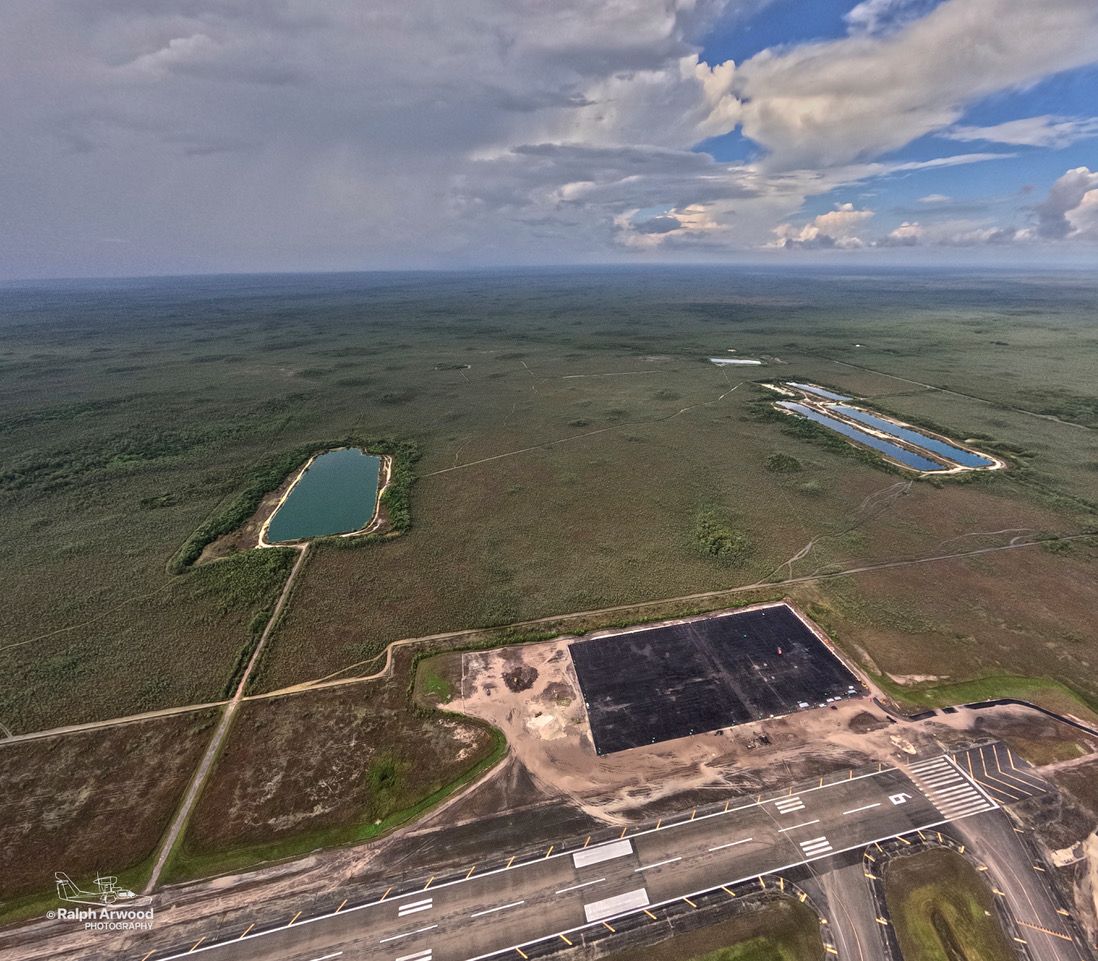 Aerial view of a runway with surrounding roads and two rectangular water bodies in a vast green plain under a cloudy sky with blue patches.