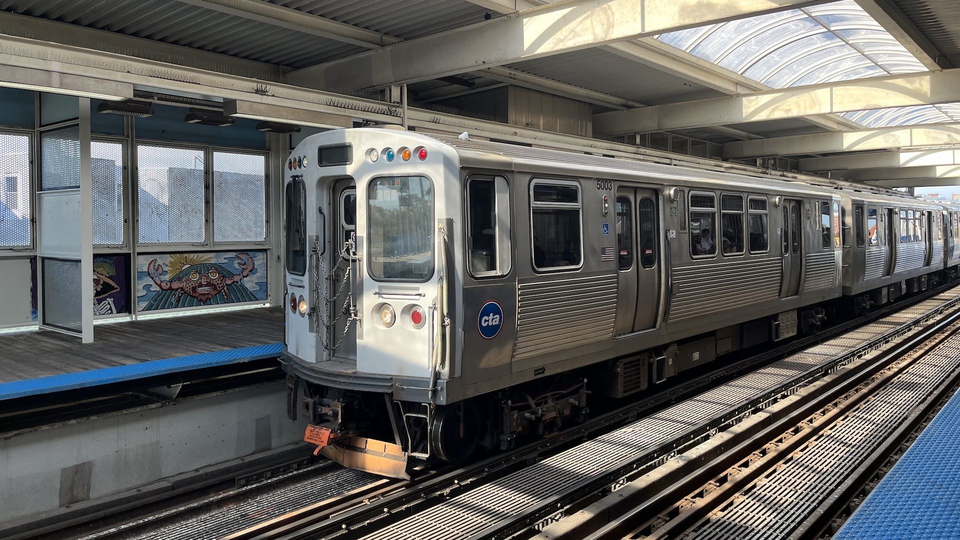 Silver Chicago CTA train number 5003 at a station with a covered platform and blue tactile paving, under a curved translucent roof, in daylight with shadows on train tracks.