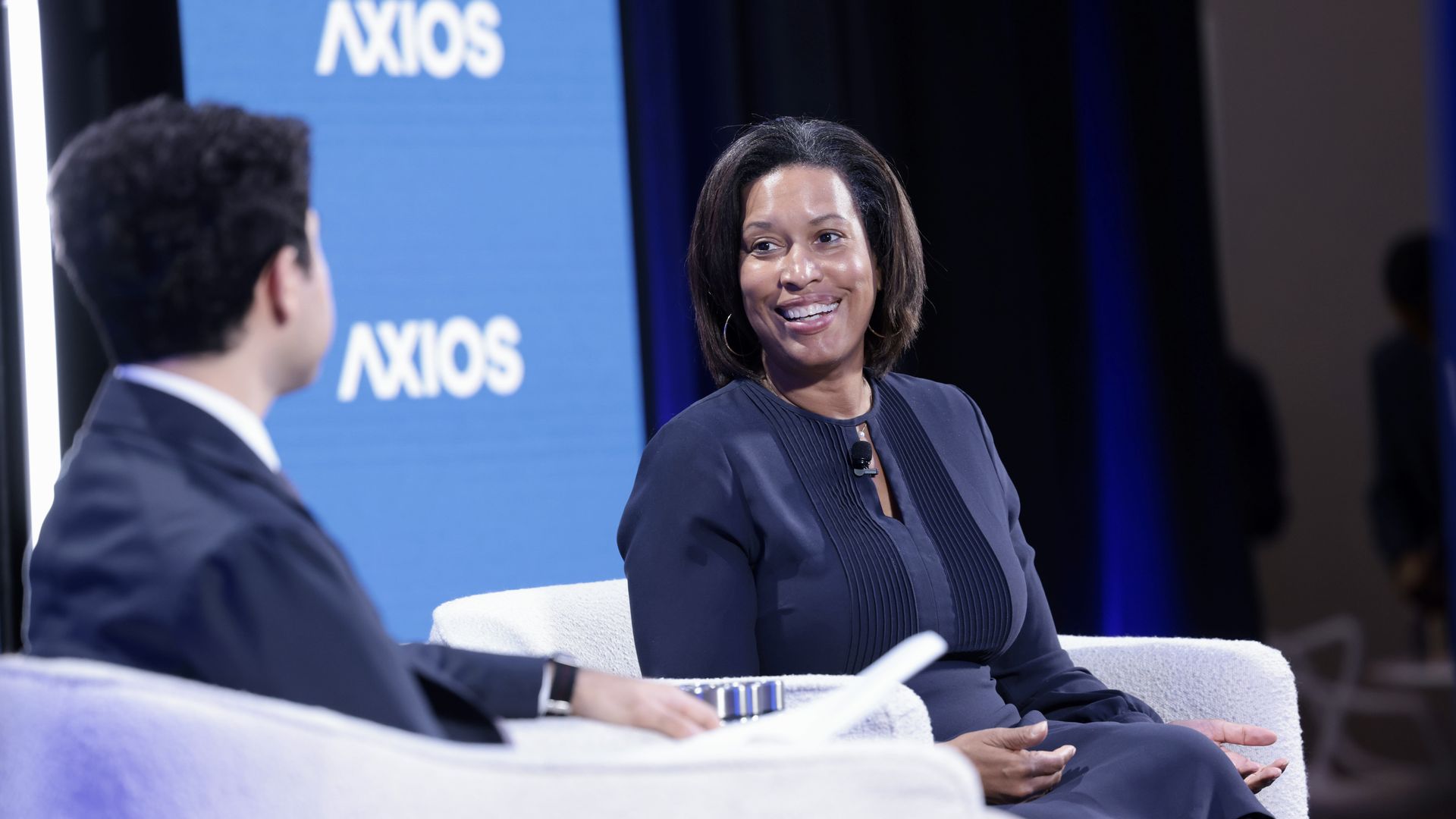 Panel discussion scene: Mayor Bowser, smiling and in a navy outfit with a microphone sits on a white chair, talking with a reporter in a suit; a blue Axios backdrop is visible behind them.