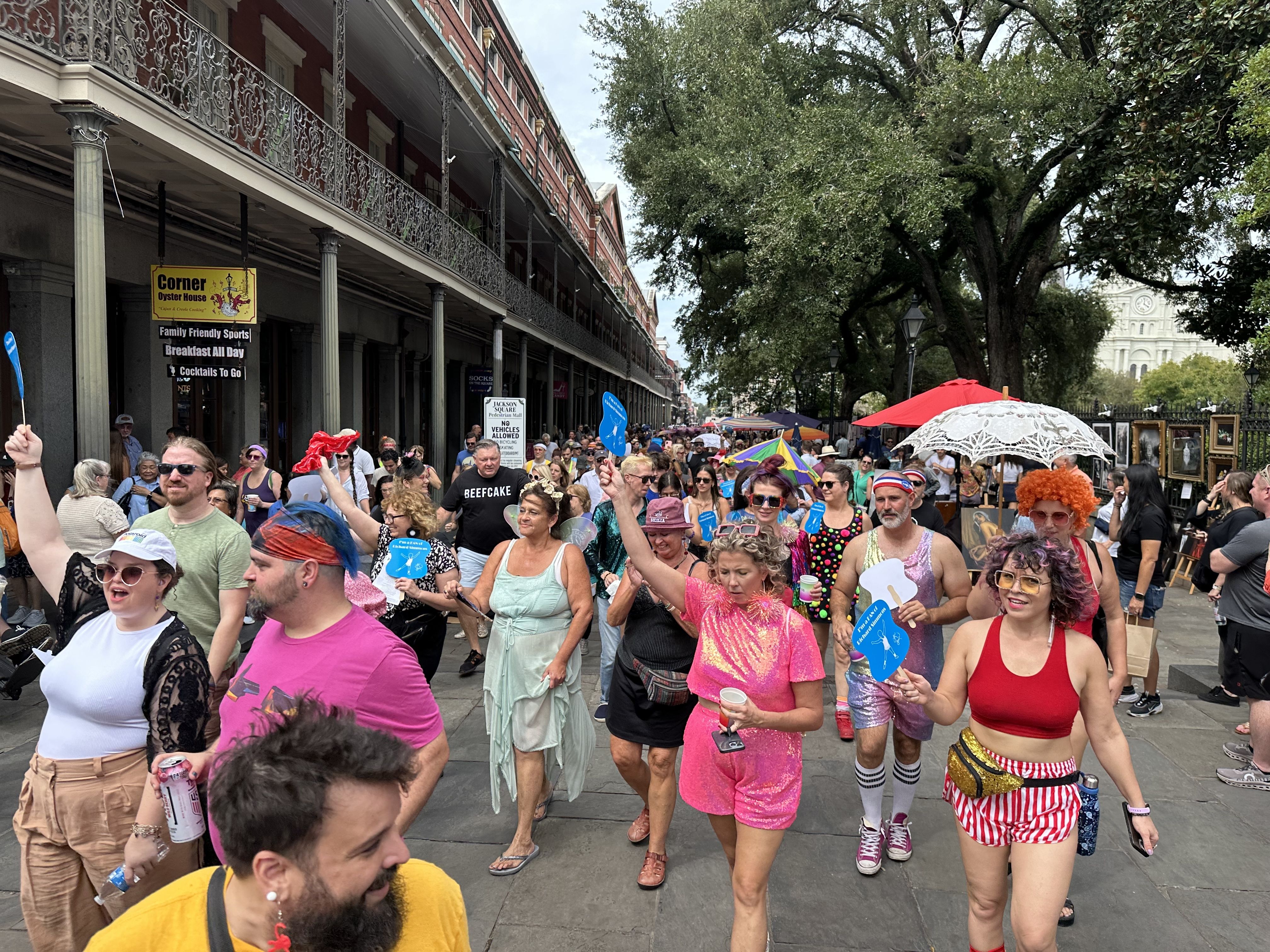 People wave to the camera as they walk past. Some are dressed in 1980s-style workout gear and wigs.