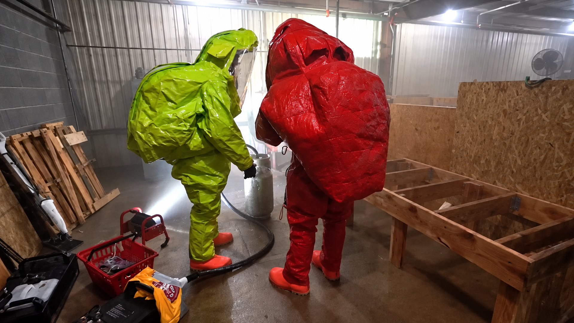 Two people in hazmat suits, one lime green and the other red, stand in a metal-walled workshop handling a gas cylinder with a hose, surrounded by pallets, plywood panels, and tools.