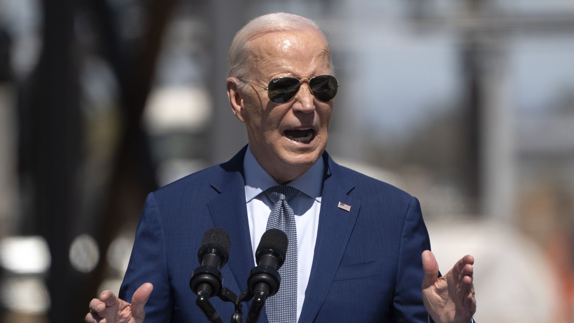 President Biden, wearing a dark blue suit, light blue shirt pale blue tie and aviator sunglasses, speaks at a podium with microphones outdoors.