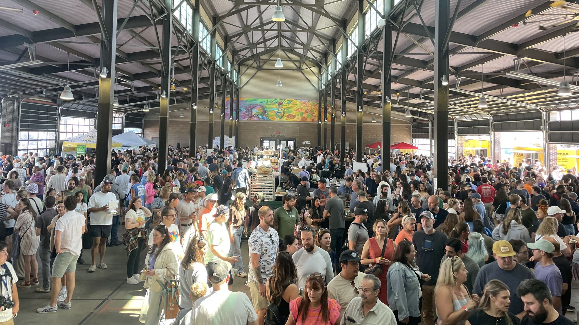 A bustling indoor market in a large warehouse with dark steel trusses, high ceiling, hanging lights, and sunlit windows. A crowded hall with people, booths, and colorful mural on the back wall.