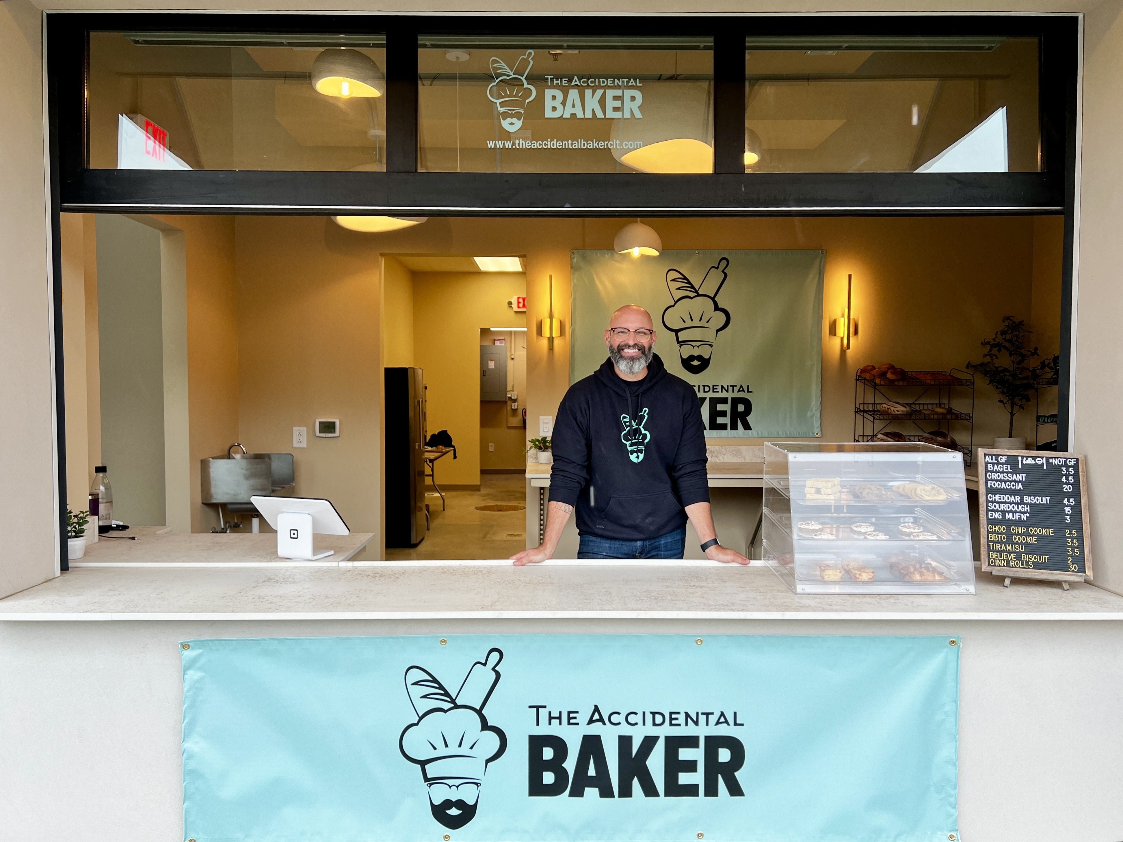 man standing behind counter with sign that reads "Accidental Baker"