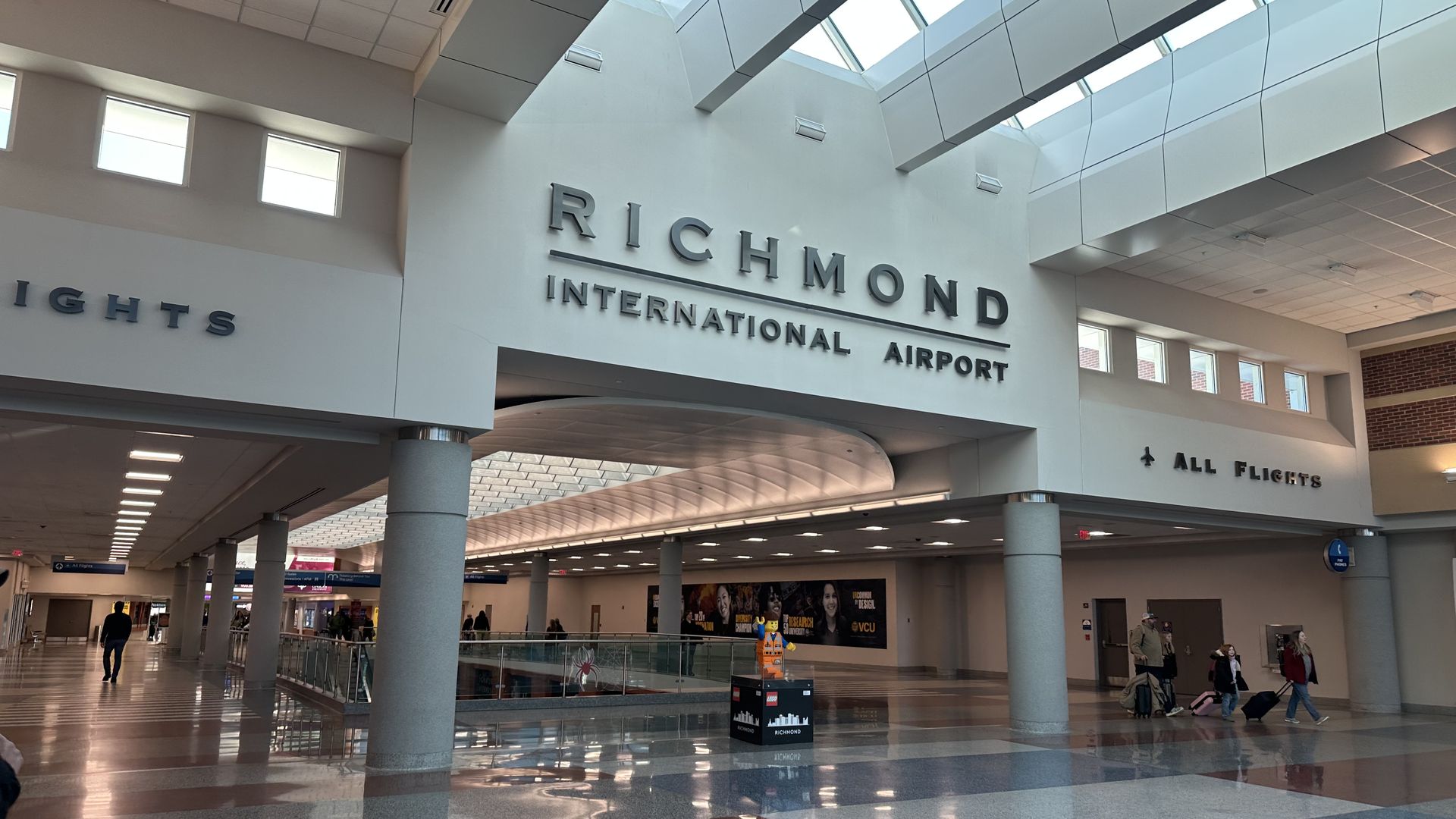 Interior of Richmond International Airport terminal with polished floors, columns, skylights, and a LEGO figure display in the center. Several travelers walk with luggage.