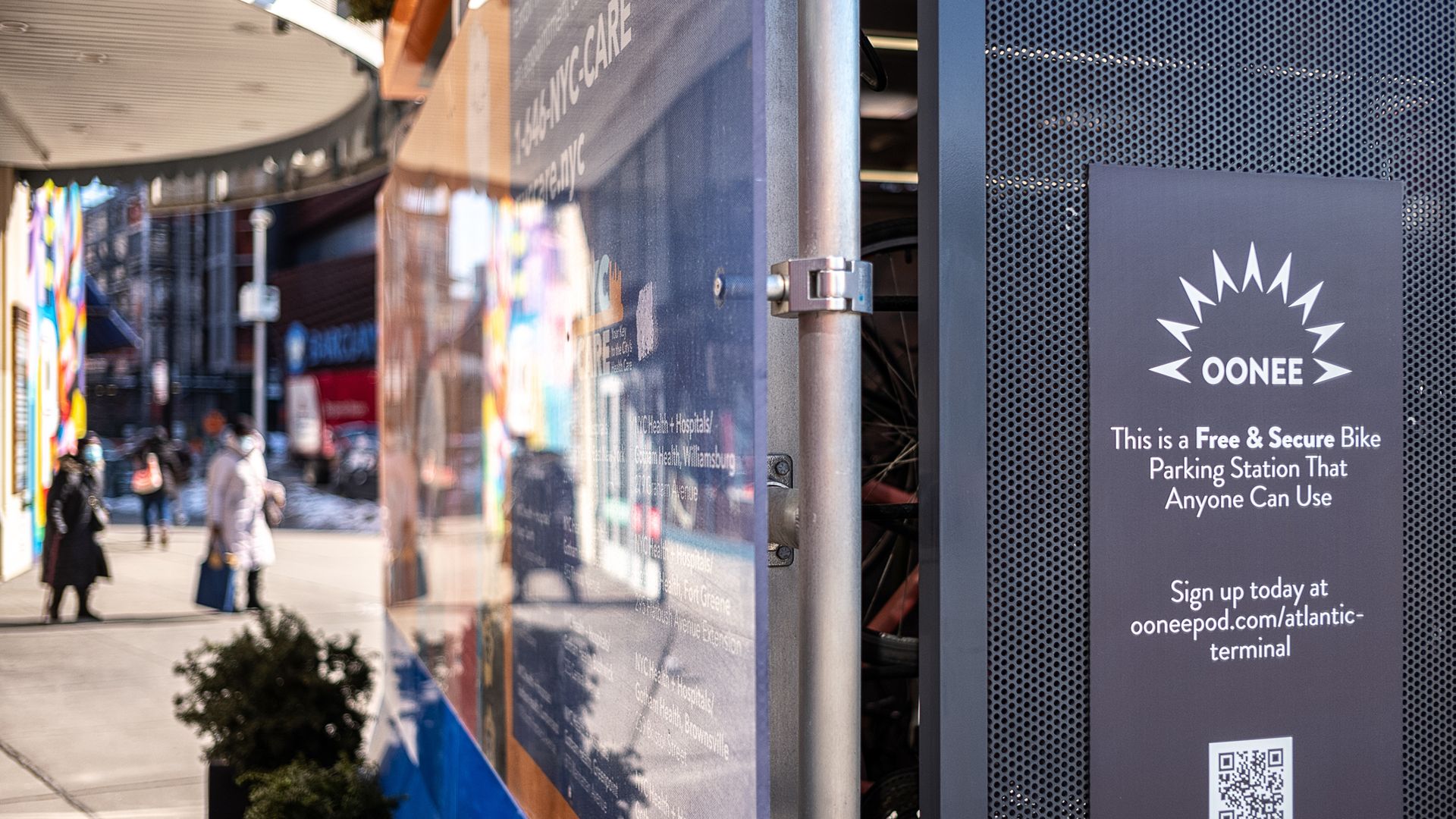 A glass-paneled shelter building on a public sidewalk with a sign on the side reading: 'Oonee, This is a Free & Secure Bike Parking Station That Anyone Can Use'