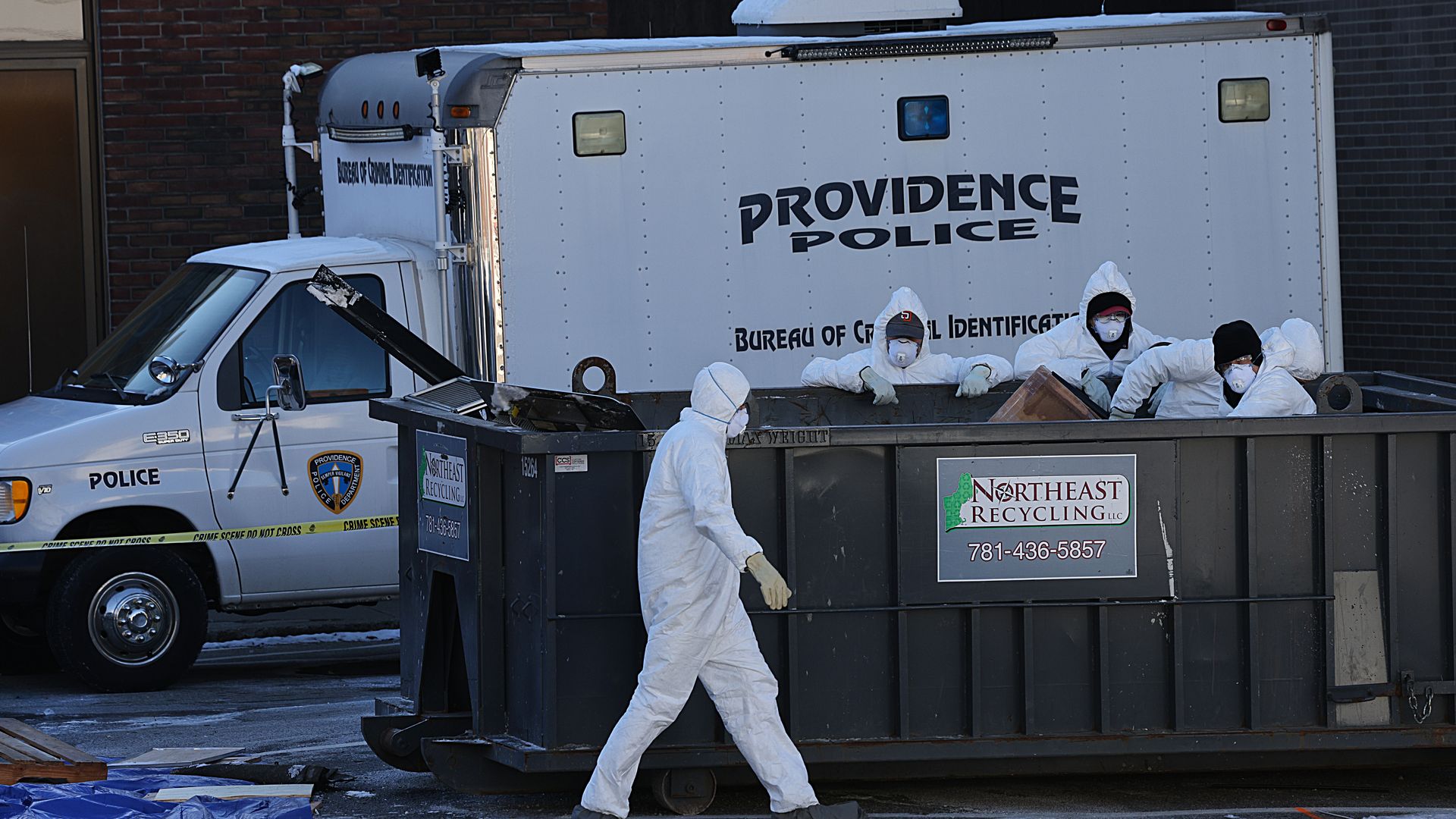 People in white protective suits and masks work around a large black dumpster labeled Northeast Recycling, beside a white Providence Police truck marked Bureau of Criminal Identification.