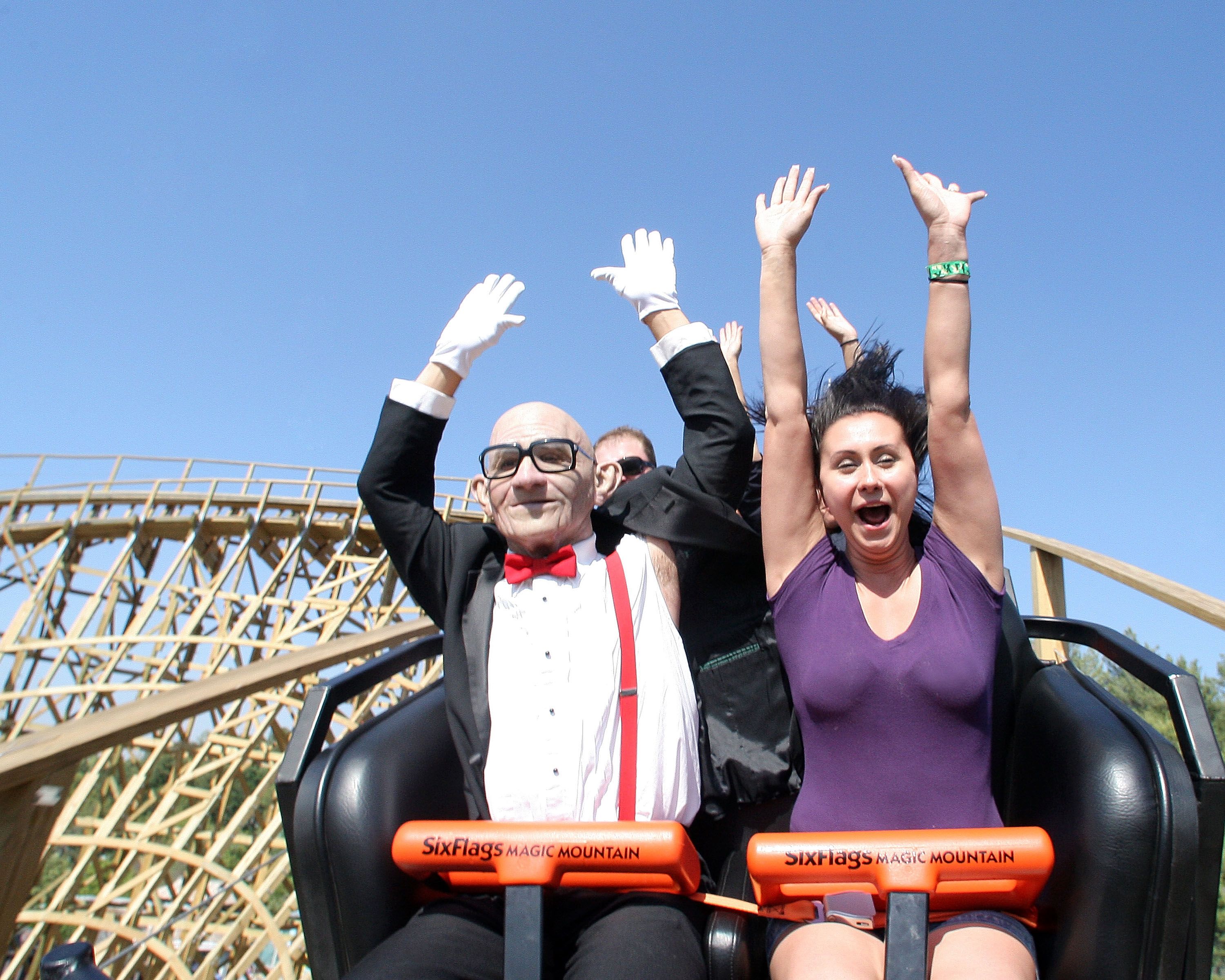 A man in a costume dressed as a much older man rides a rollercoaster next to another younger person. They have their hands in the air.