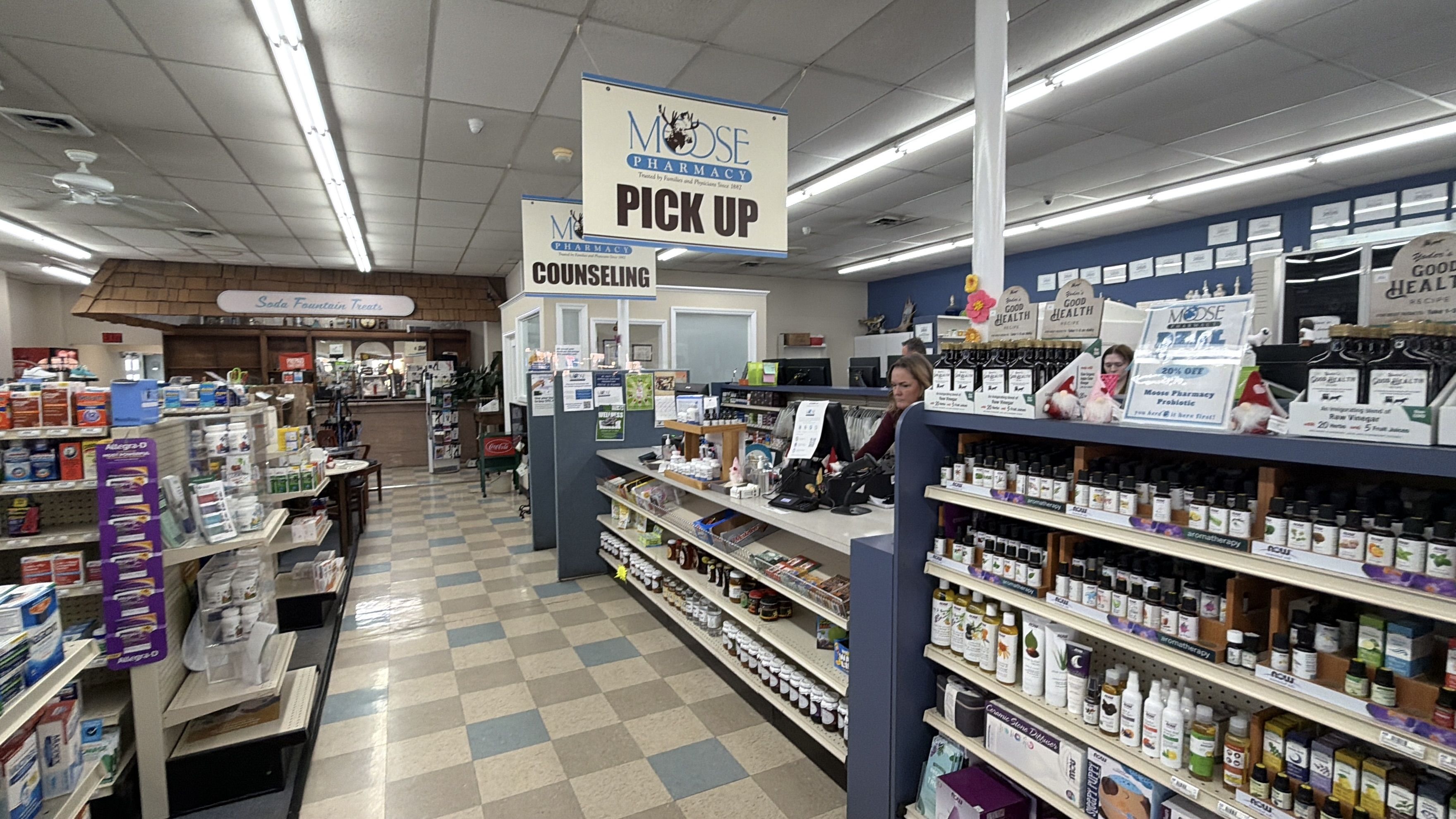 Interior of Moose Pharmacy with counters labeled Pick Up and Counseling, shelves stocked with medicines and supplements, and a Soda Fountain Treats sign in the background.