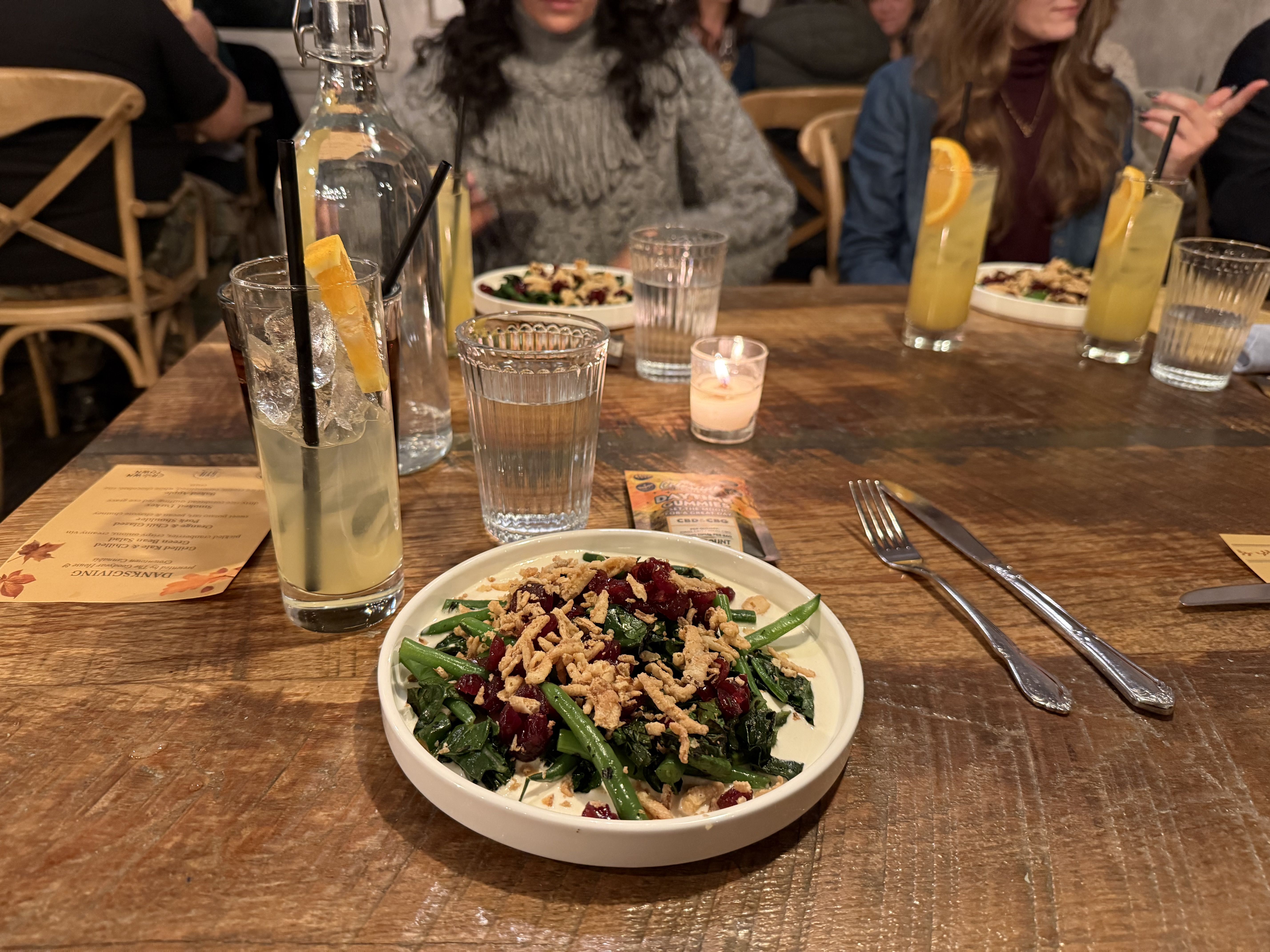 Dining table with a vegetable salad in a white bowl topped with dried cranberries and crispy bits, surrounded by glasses of water and yellow cocktails with orange slices, candle, and people in background.