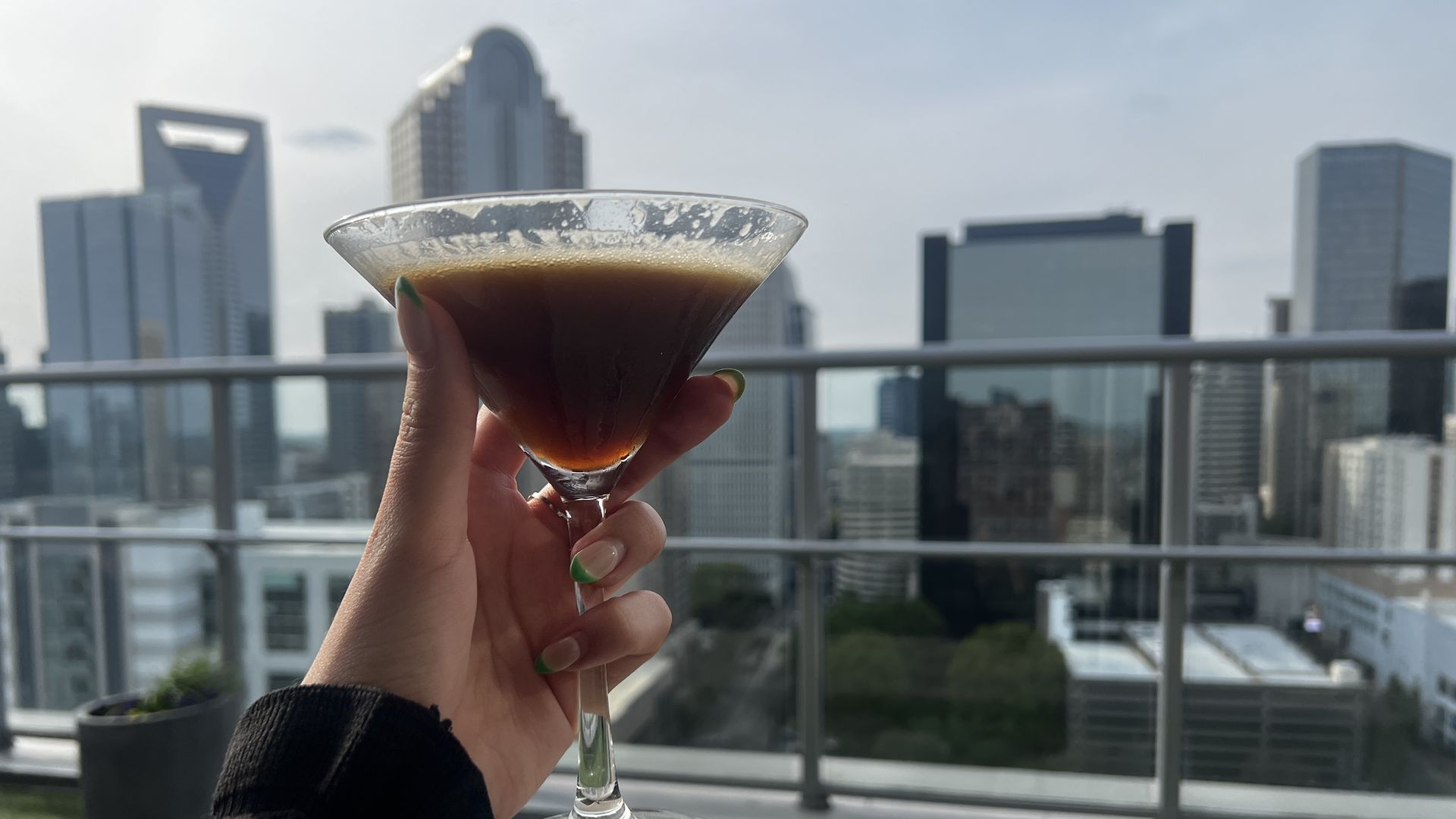 Hand with green nails holds a coupe glass of dark, layered cocktail on a balcony; a city skyline of tall buildings looms in the background.