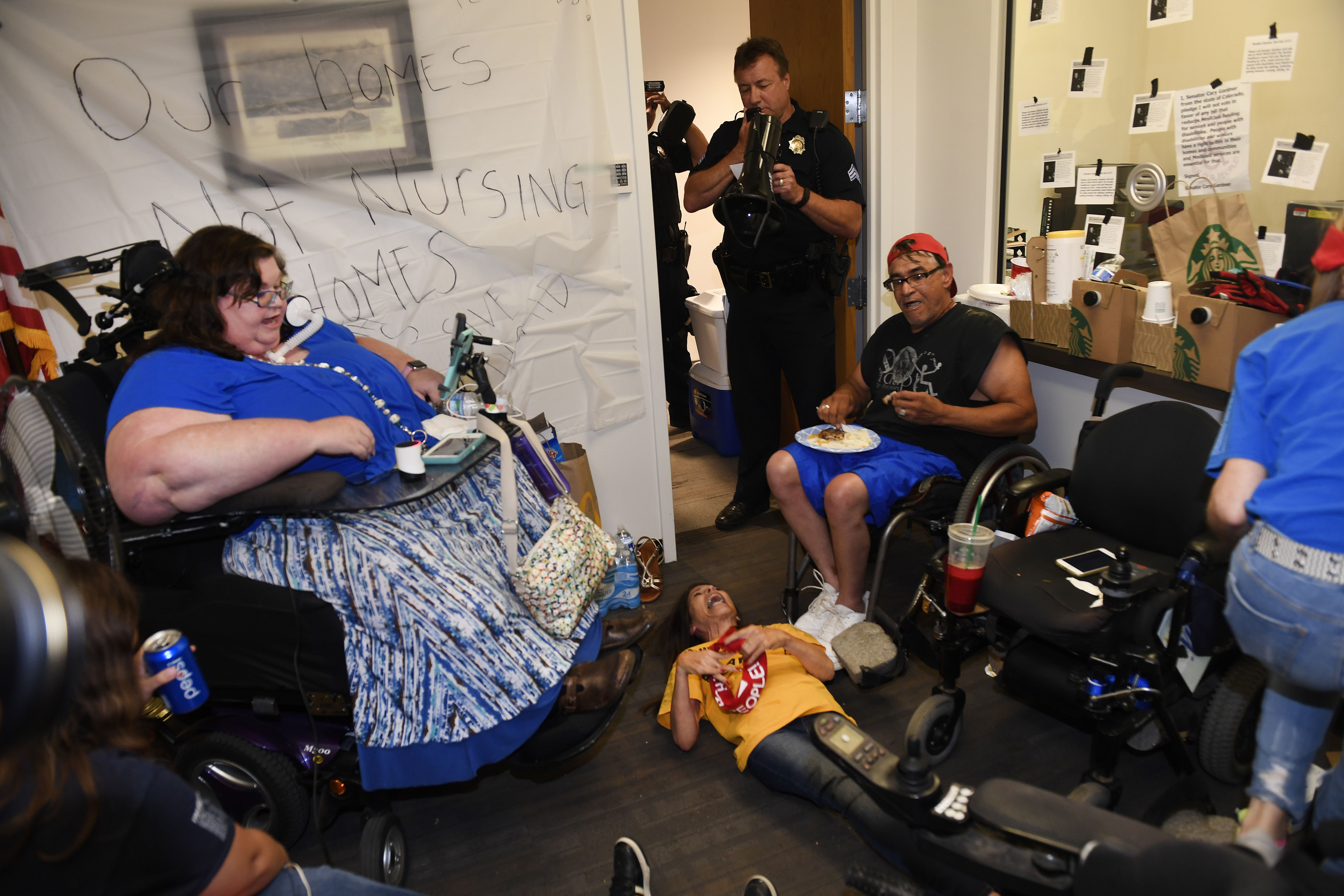 A group of people in wheelchairs, with one person lying on the ground, sits in a room surrounded by police.