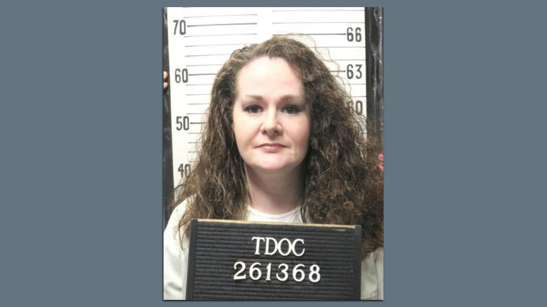 Mugshot of a woman with long curly brown hair, standing before a height chart and holding a sign that reads TDOC 261368; gray background.