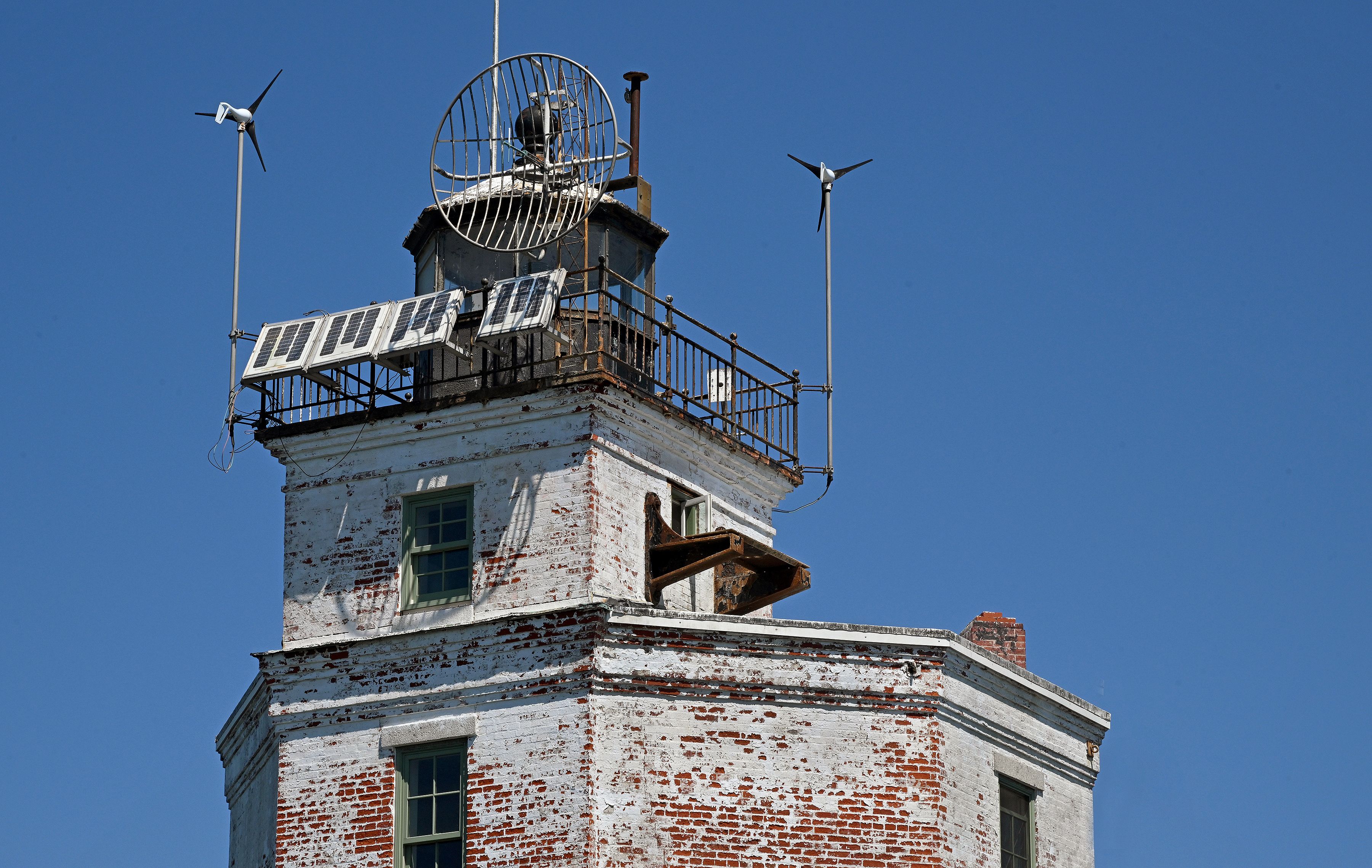 Top of an old white brick lighthouse with peeling paint, featuring solar panels, antennas, and two small wind turbines against a clear blue sky.