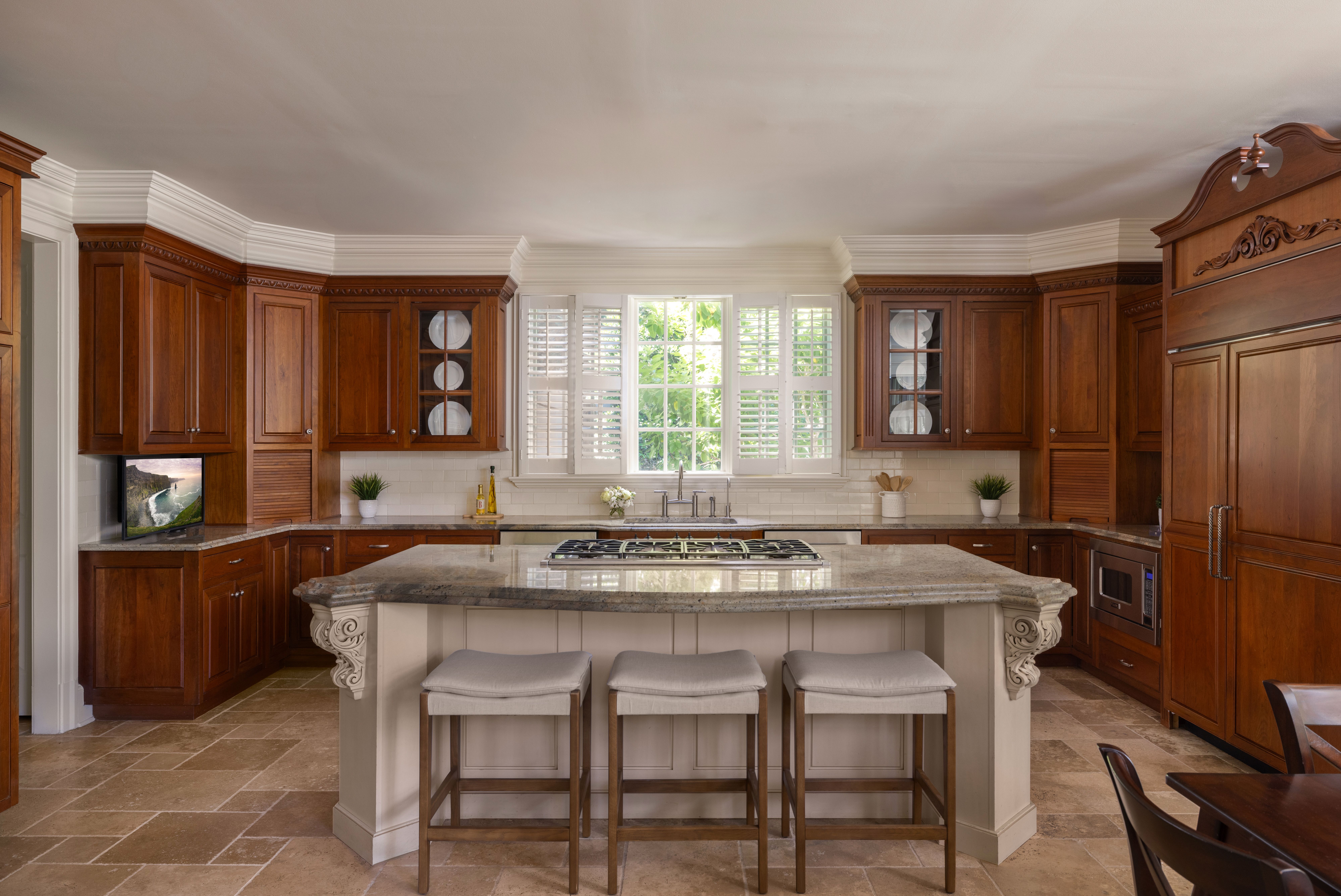 Bright kitchen with dark wood cabinets, white crown molding, and a granite island with four stools. A window wall with white shutters overlooks greenery; sink sits beneath the windows.