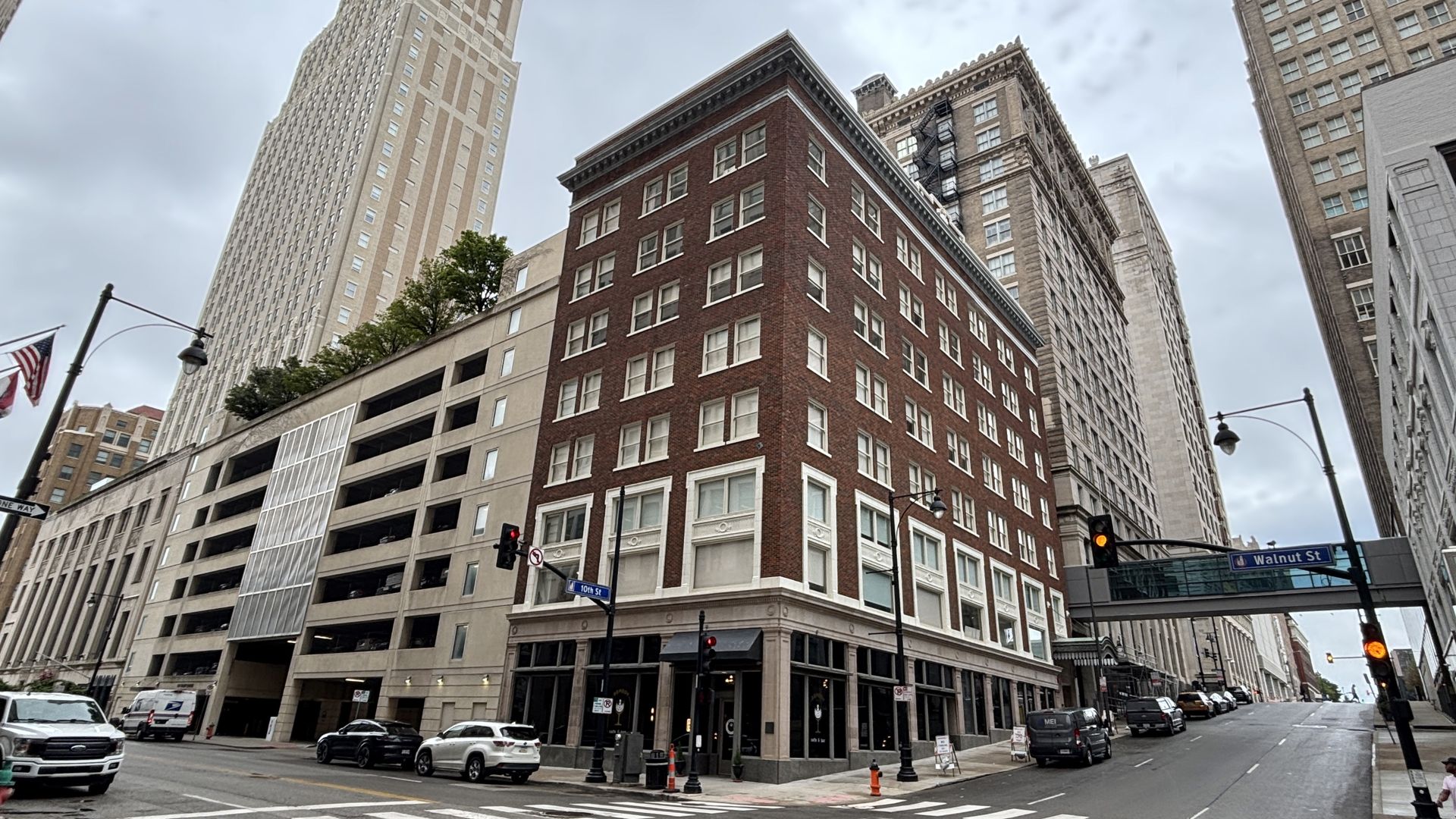 Urban street corner with a red brick four-story building at the center, flanked by tall beige and gray towers; cars parked along the curb, an American flag, and an overcast sky.