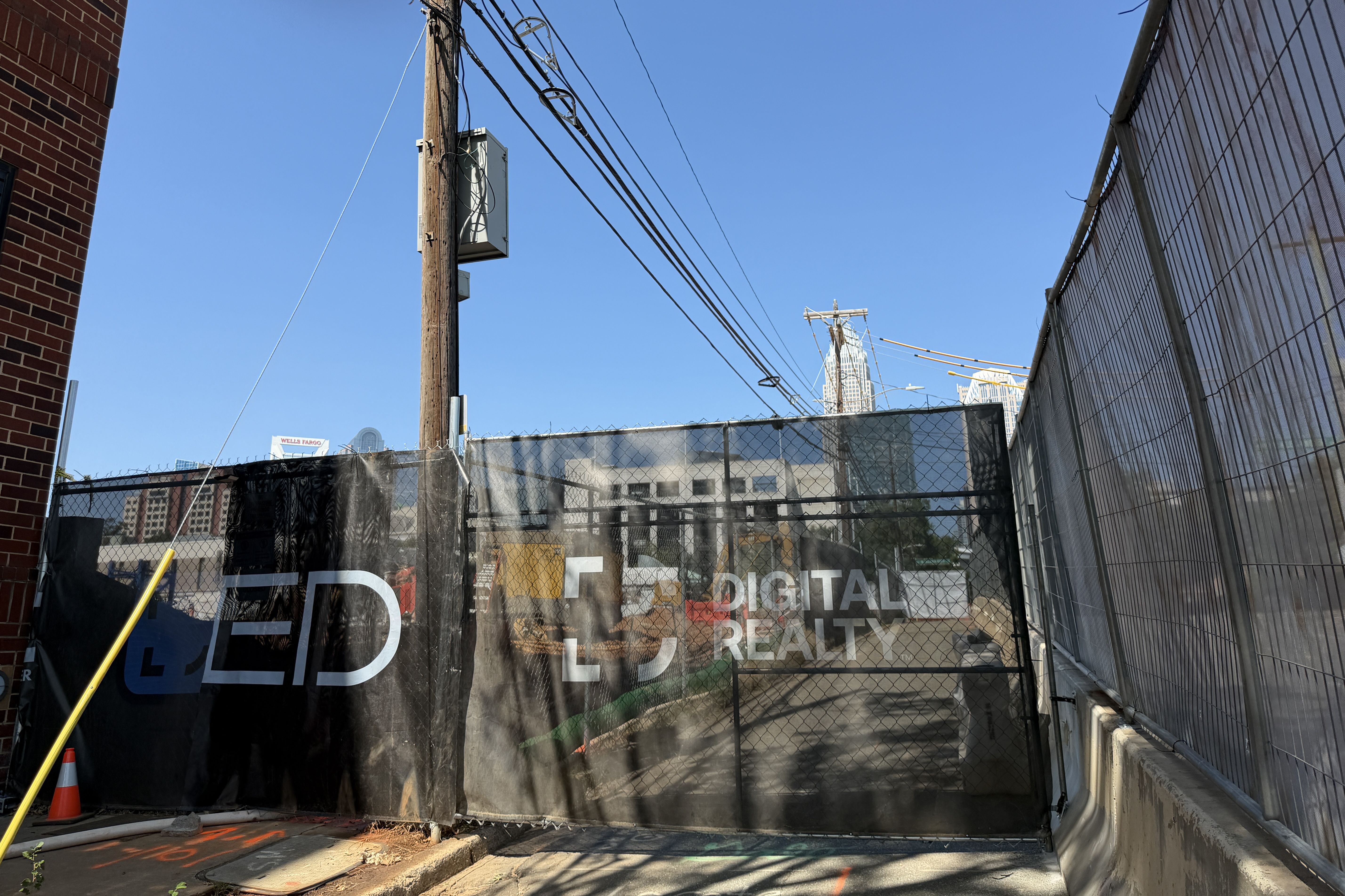 Urban alley with a tall wooden utility pole and overhead power lines, brick wall on the left, corrugated fence on the right, and distant skyscrapers beneath a clear blue sky.