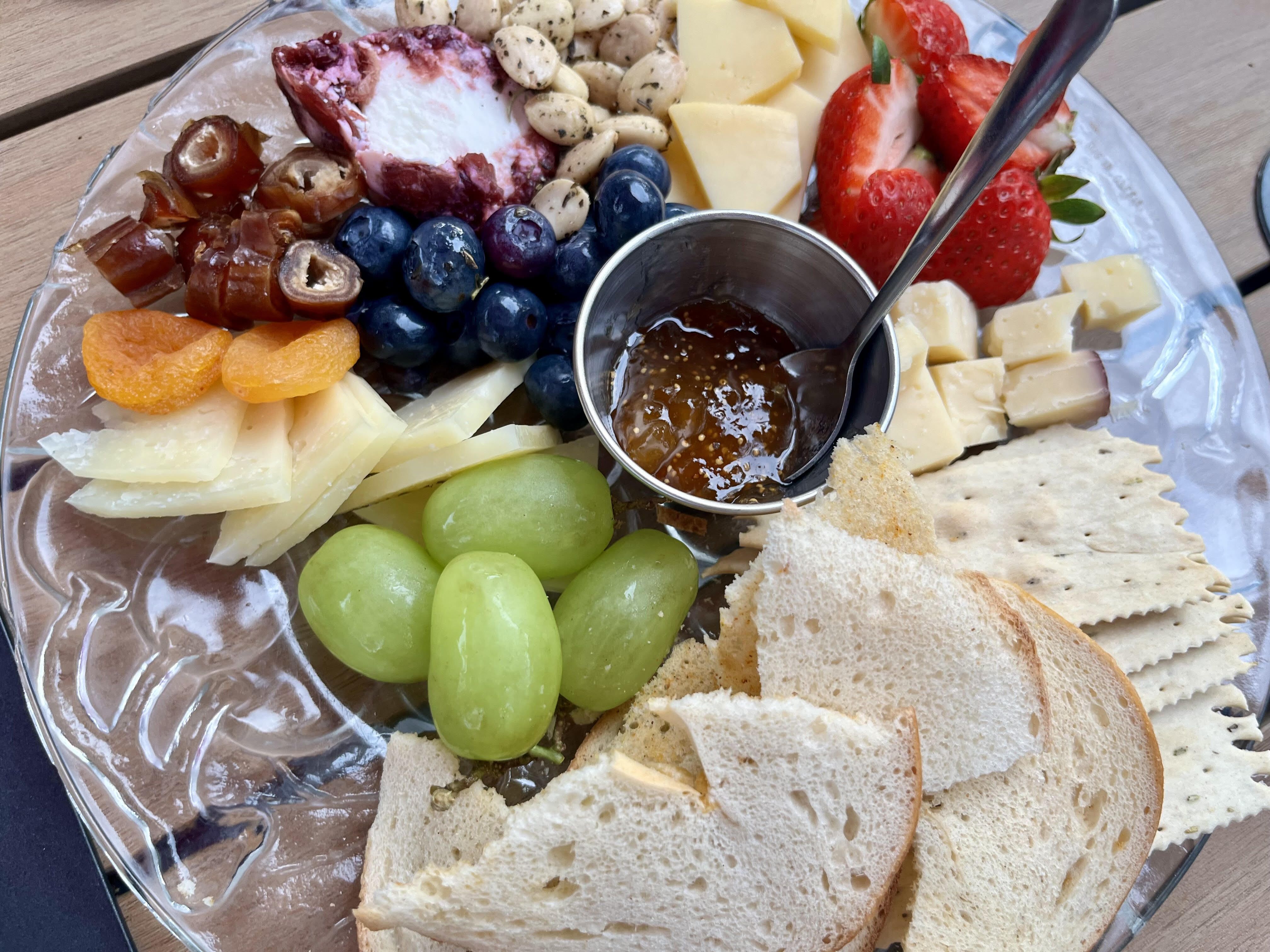 Cheese and fruit platter with assorted cheeses, green grapes, blueberries, strawberries, dried apricots, dates, herbed nuts, crackers, sliced bread, and a small metal cup of jam with a spoon.