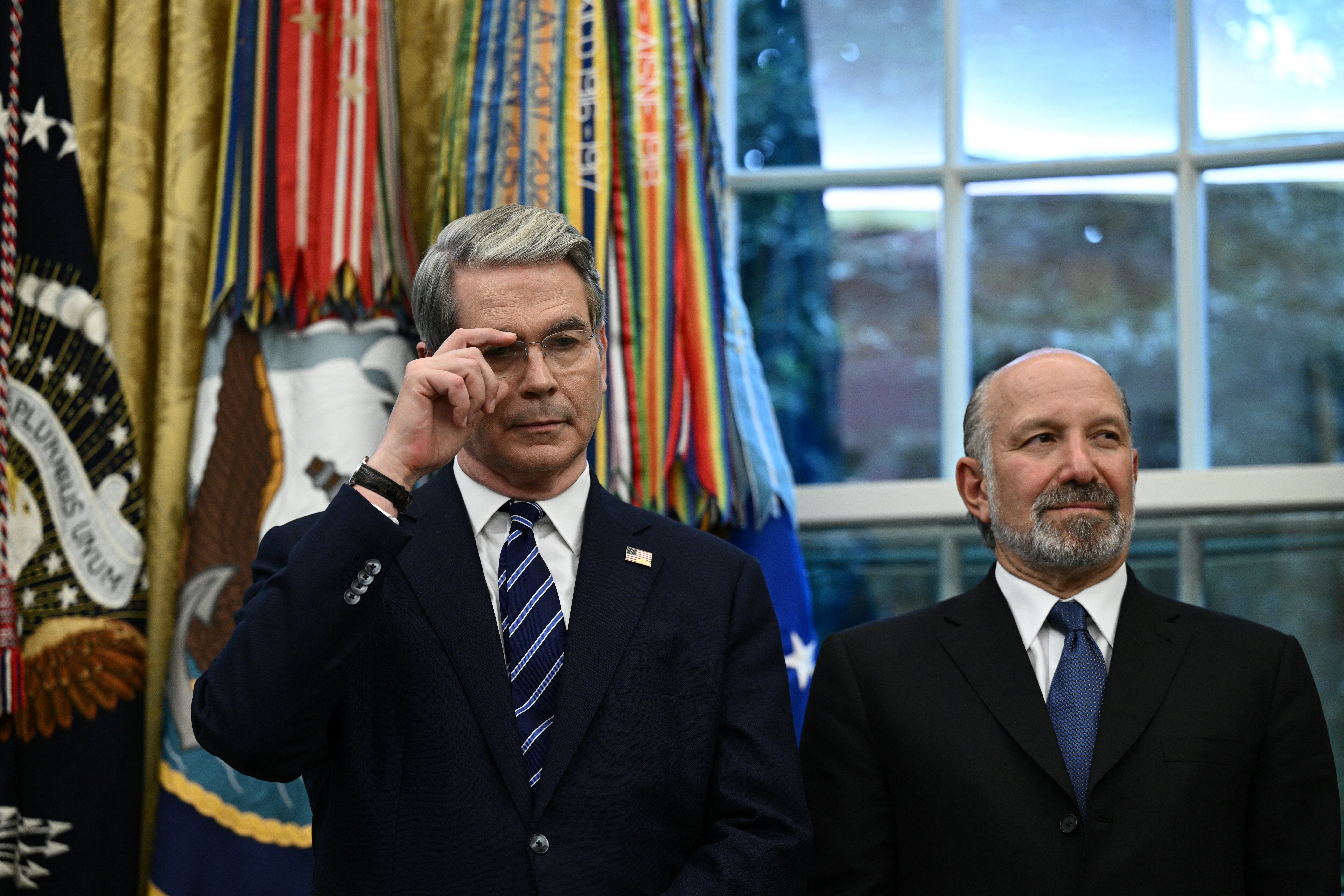 Two men in dark suits and ties stand in front of colorful military ribbons and a window; the man on the left adjusts his glasses and has gray hair, the man on the right has a beard.