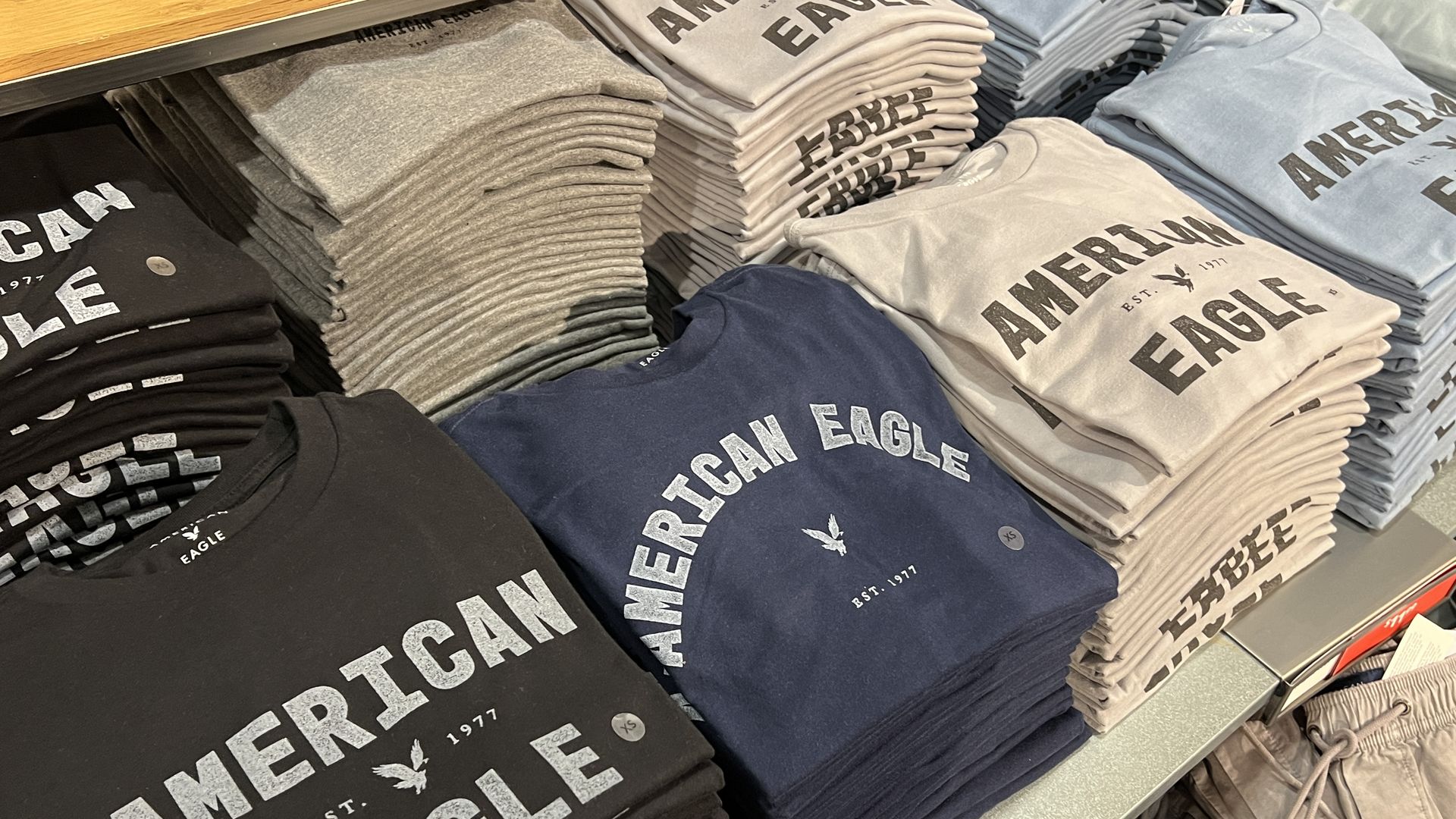 Stacks of folded American Eagle T-shirts in black, navy blue, light gray, and light blue on a store shelf.