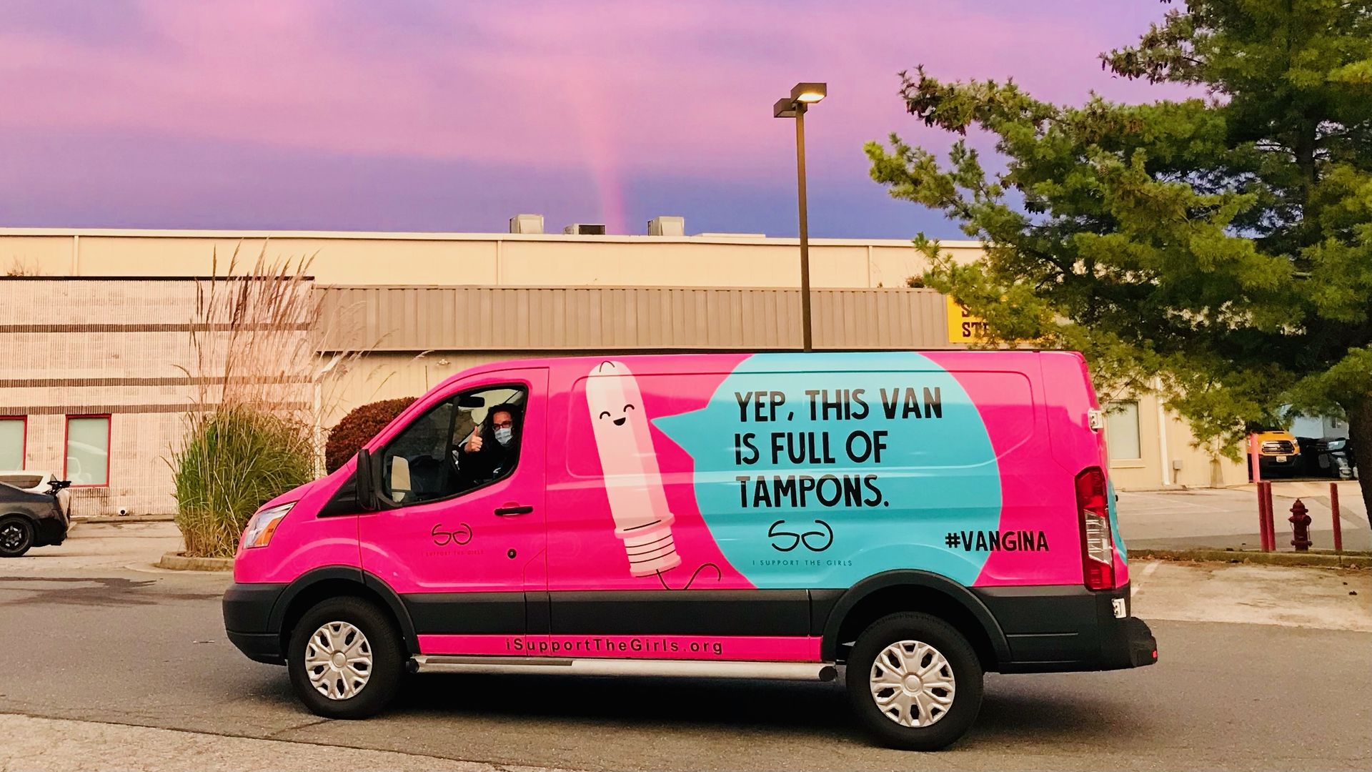 A person wearing a face mask gives a thumbs' up in the driver's seat of a bright pink van with cartoon tampon on the side with a speech bubble reading "Yep, this van is full of tampons"