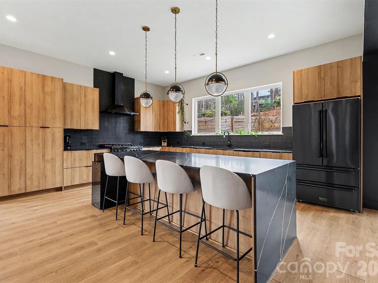 Modern kitchen with wood cabinets, black backsplash, black appliances, a black marble island with white veins, four gray bar stools, wooden floor, and three spherical pendant lights.