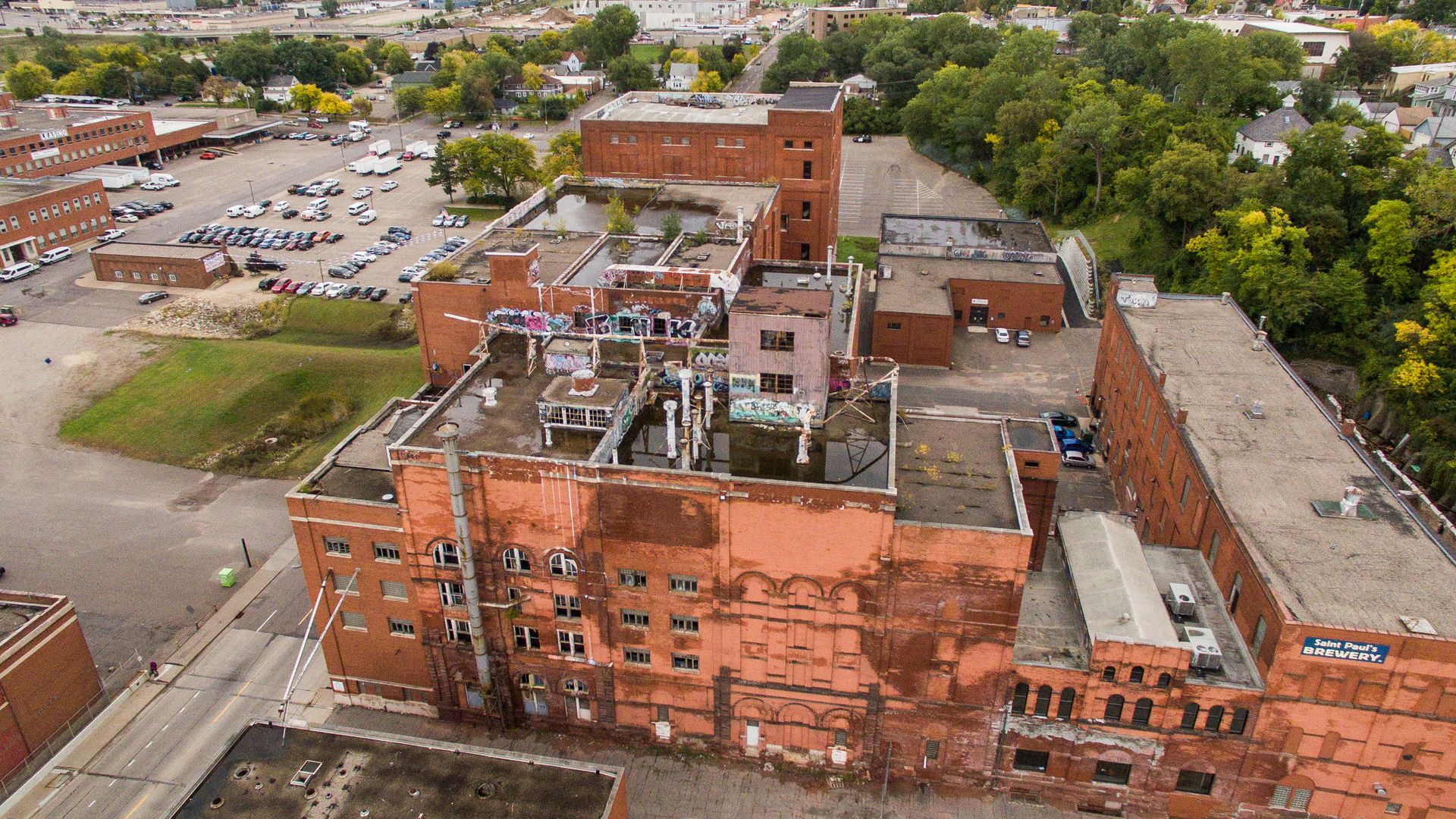 Aerial view of old brick industrial buildings with graffiti, surrounded by parking lots, greenery, and some trees, under a cloudy sky in an urban area.