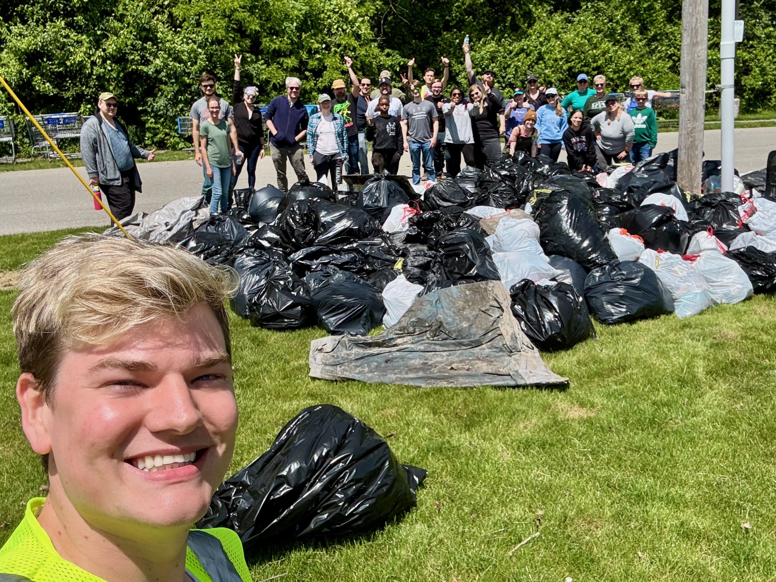 Group of people posing behind a large pile of black and white garbage bags on green grass on a sunny day, with trees and a road in the background.