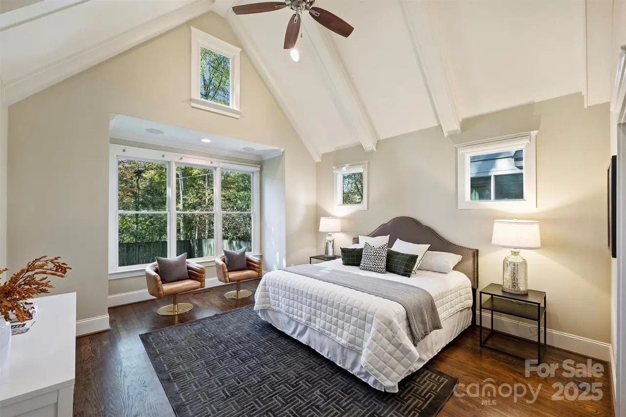 Bright bedroom with vaulted ceiling, white bedspread, gray and green pillows, two leather chairs by large windows, dark rug on wooden floor, bedside tables with lamps.