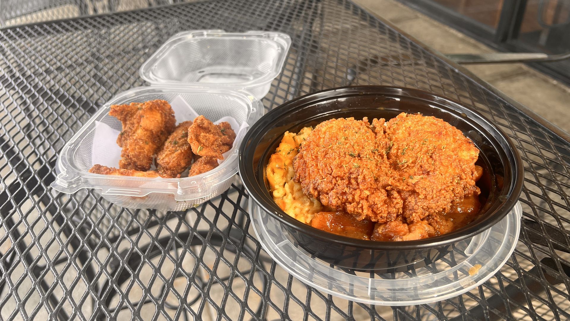 Outdoor metal mesh table with takeout: a black bowl of rice topped with crispy fried chicken and curry on the right, and a clear plastic container of fried chicken pieces on the left.