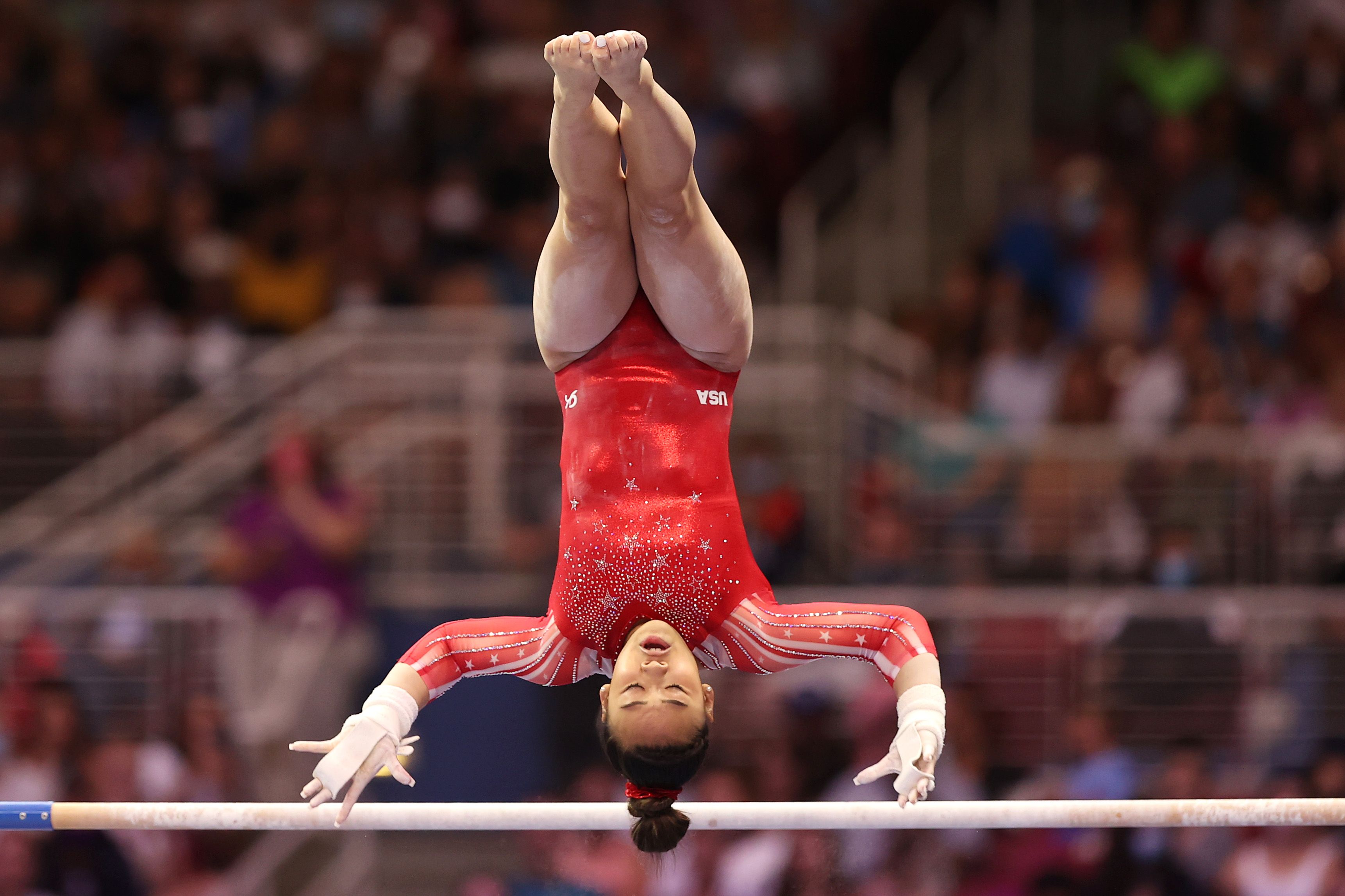 Photo of Suni Lee swinging in the air in a red uniform upside down over a bar