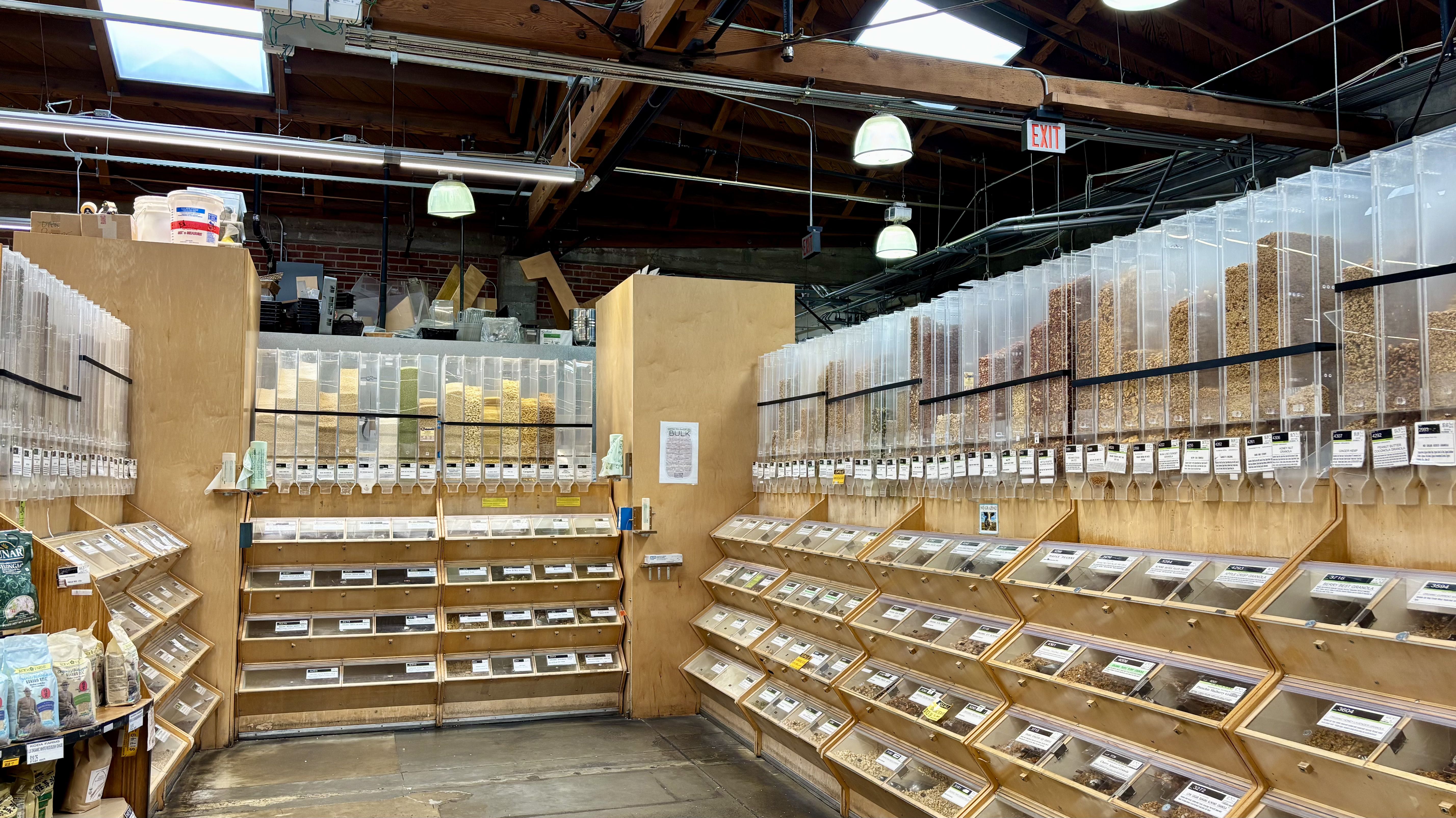 Interior of a bulk food store with wooden shelves and clear dispensers filled with various grains, nuts, and seeds under bright hanging lights and exposed wooden beams.