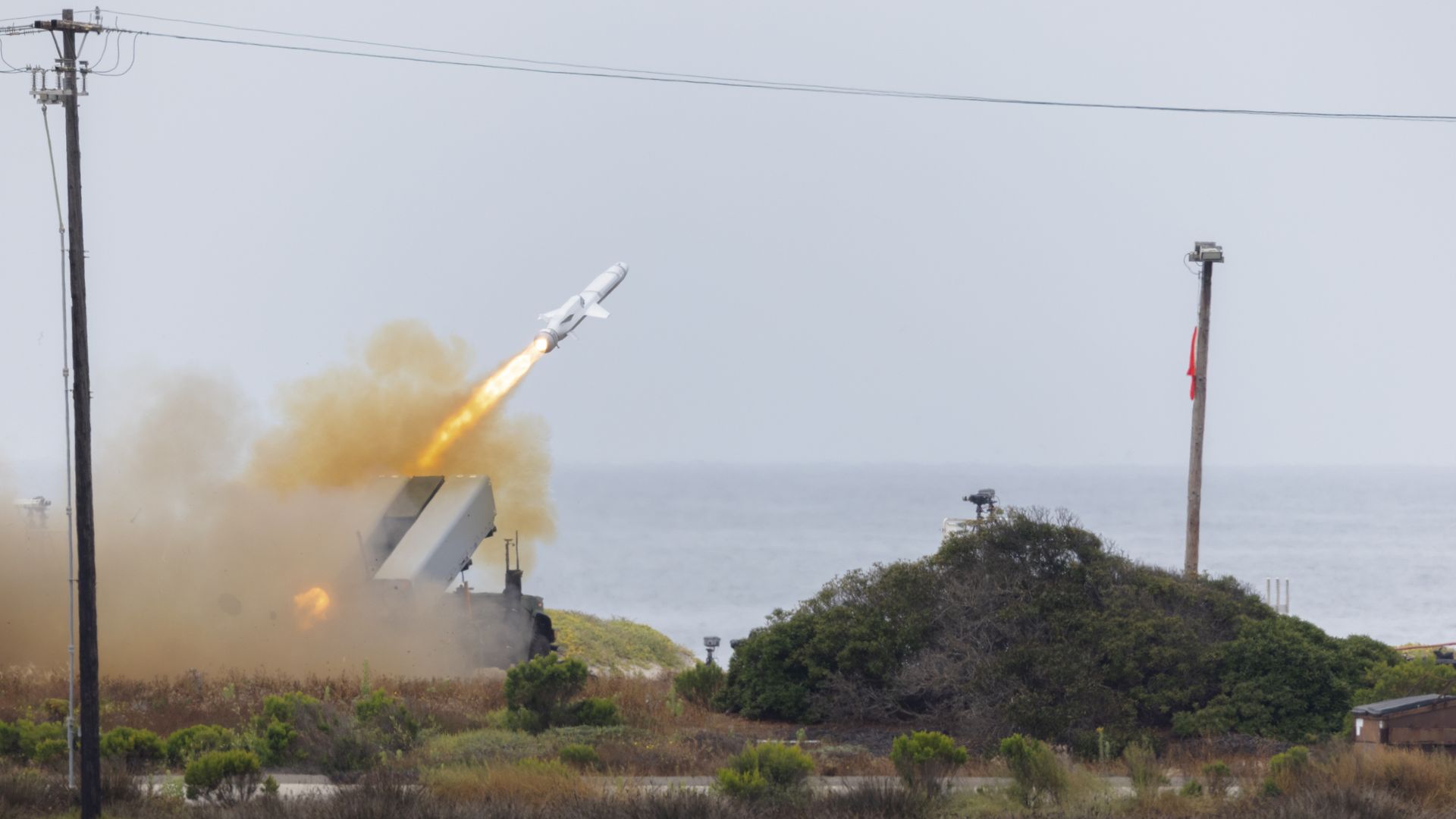 A missile launches from an unmanned vehicle near the waterfront. Telephone poles are seen on either side of the photo.