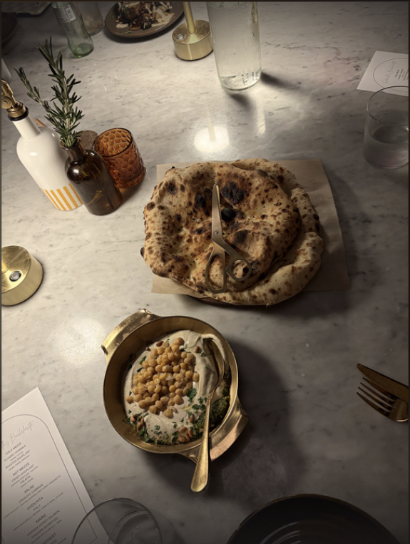 Overhead view of a marble table with a large naan on a wooden board, scissors resting on it. In front, a brass dish of hummus topped with chickpeas and herbs; rosemary sprig and bottles in background.