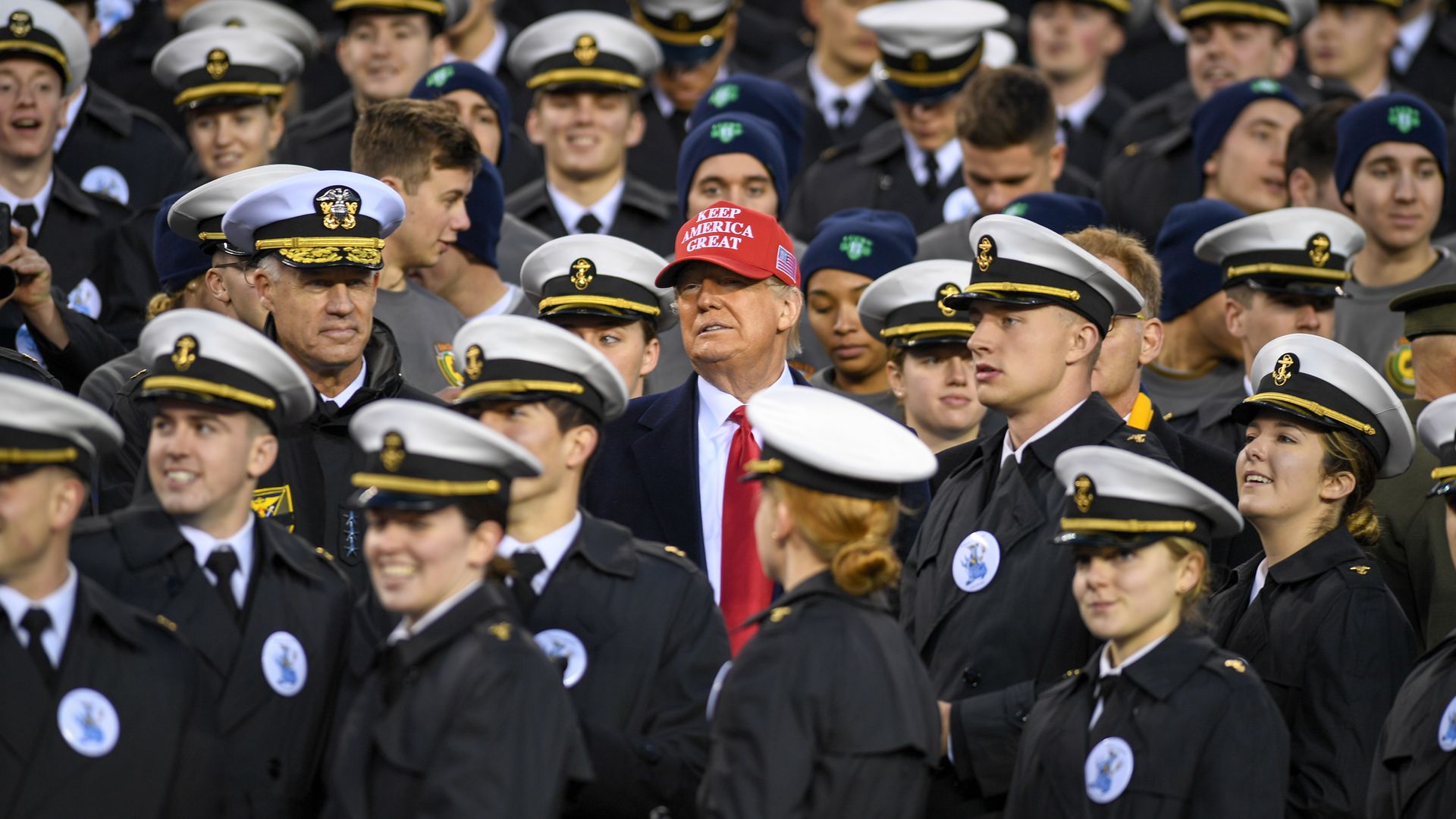 Donald Trump stands amid a sea of Naval Academy cadets. He is wearing a bright red MAGA hat.