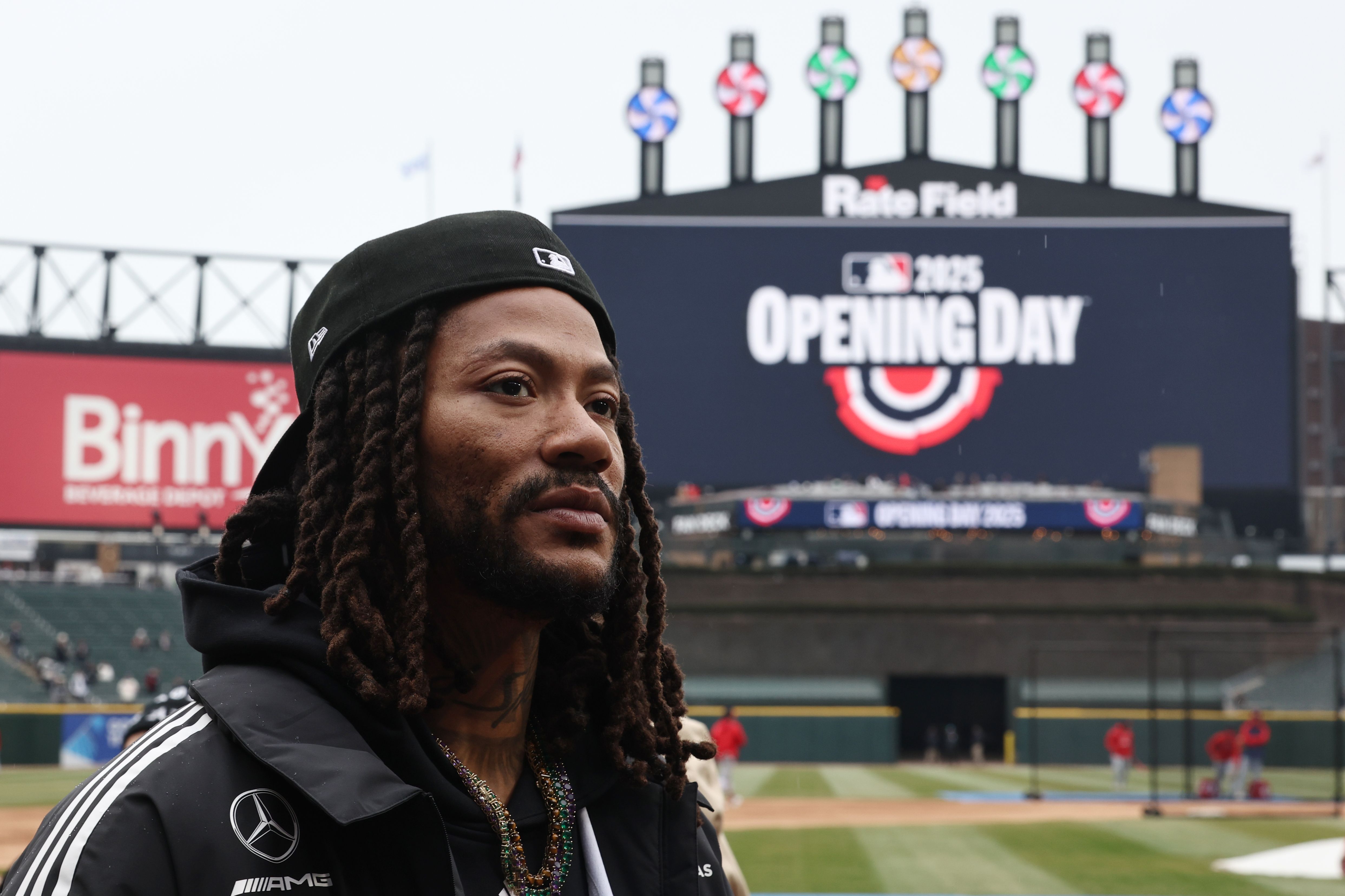Man with dreadlocks wearing a black cap and jacket at a baseball stadium with a scoreboard showing "MLB 2025 Opening Day" and colorful lights at the top.