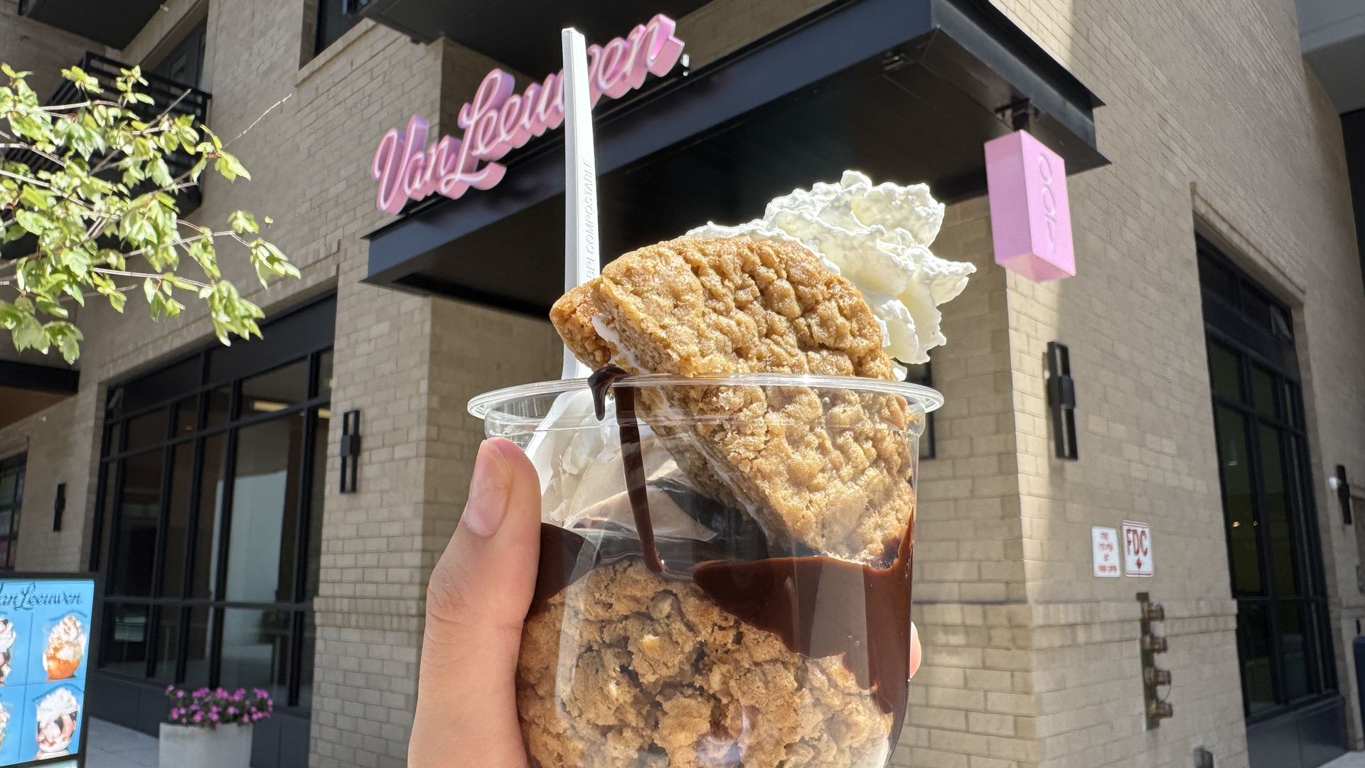 Hand holding a clear plastic cup with whipped cream, chocolate sauce, and two oatmeal cookies in front of a beige brick building with a pink "Van Leeuwen" sign.
