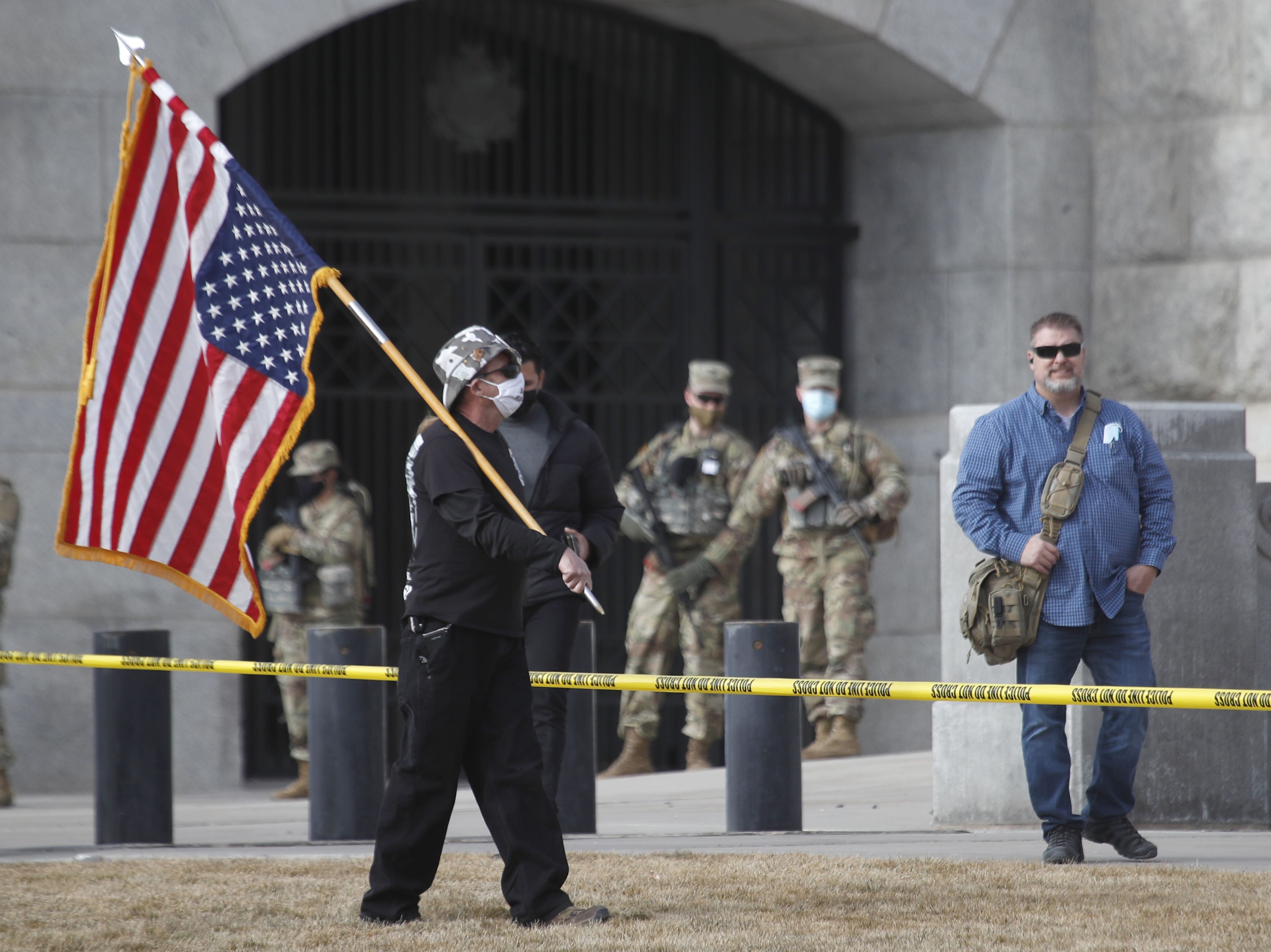 Members of the Utah National Guard stand watch as a man carries an upside down American flag as he walks the grounds of the Utah State Capitol building on January 17