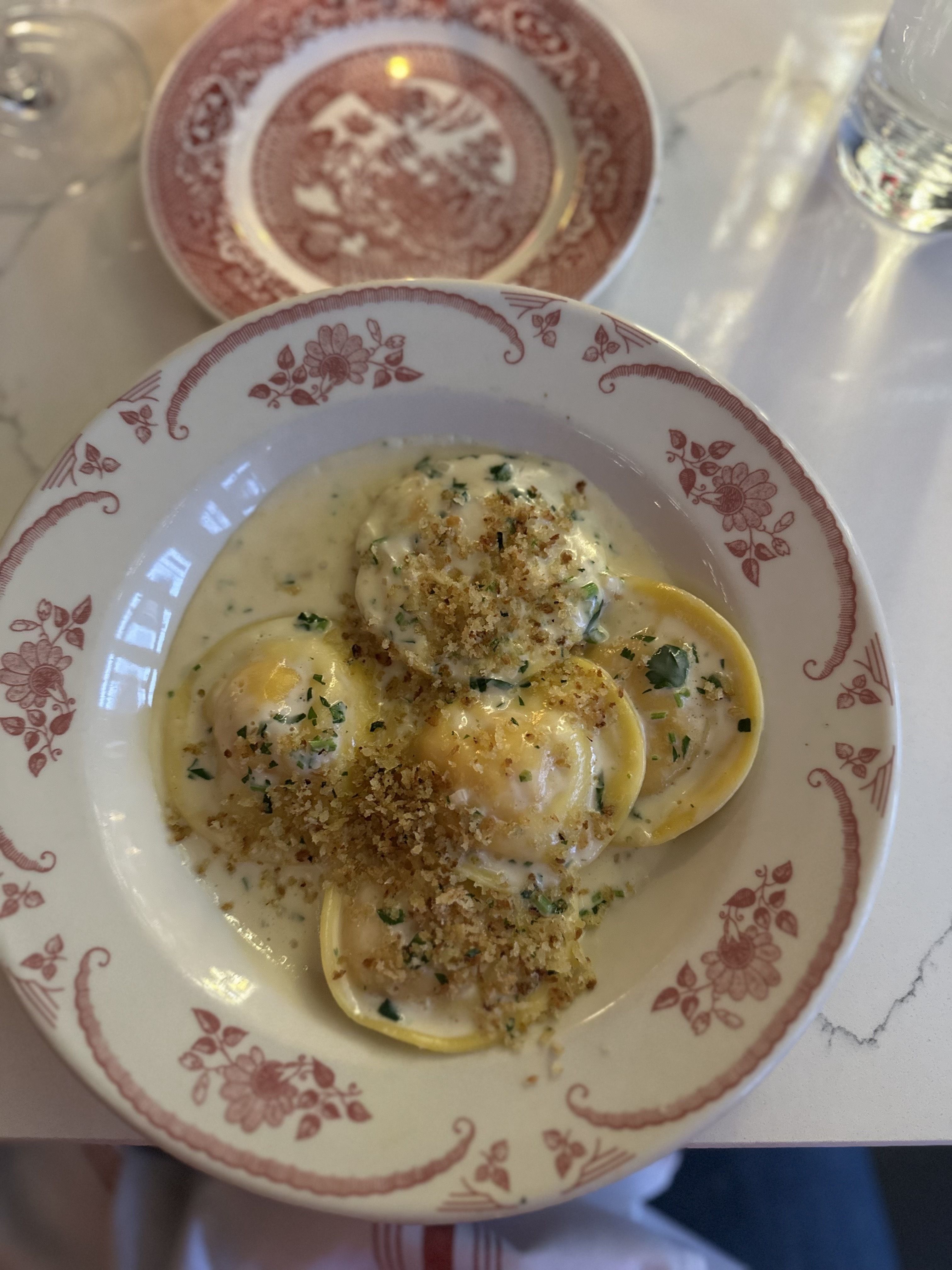 Top-down view: creamy herb ravioli on a white plate with a red floral border, topped with breadcrumbs; a second patterned plate and a glass of water sit on a light marble table.
