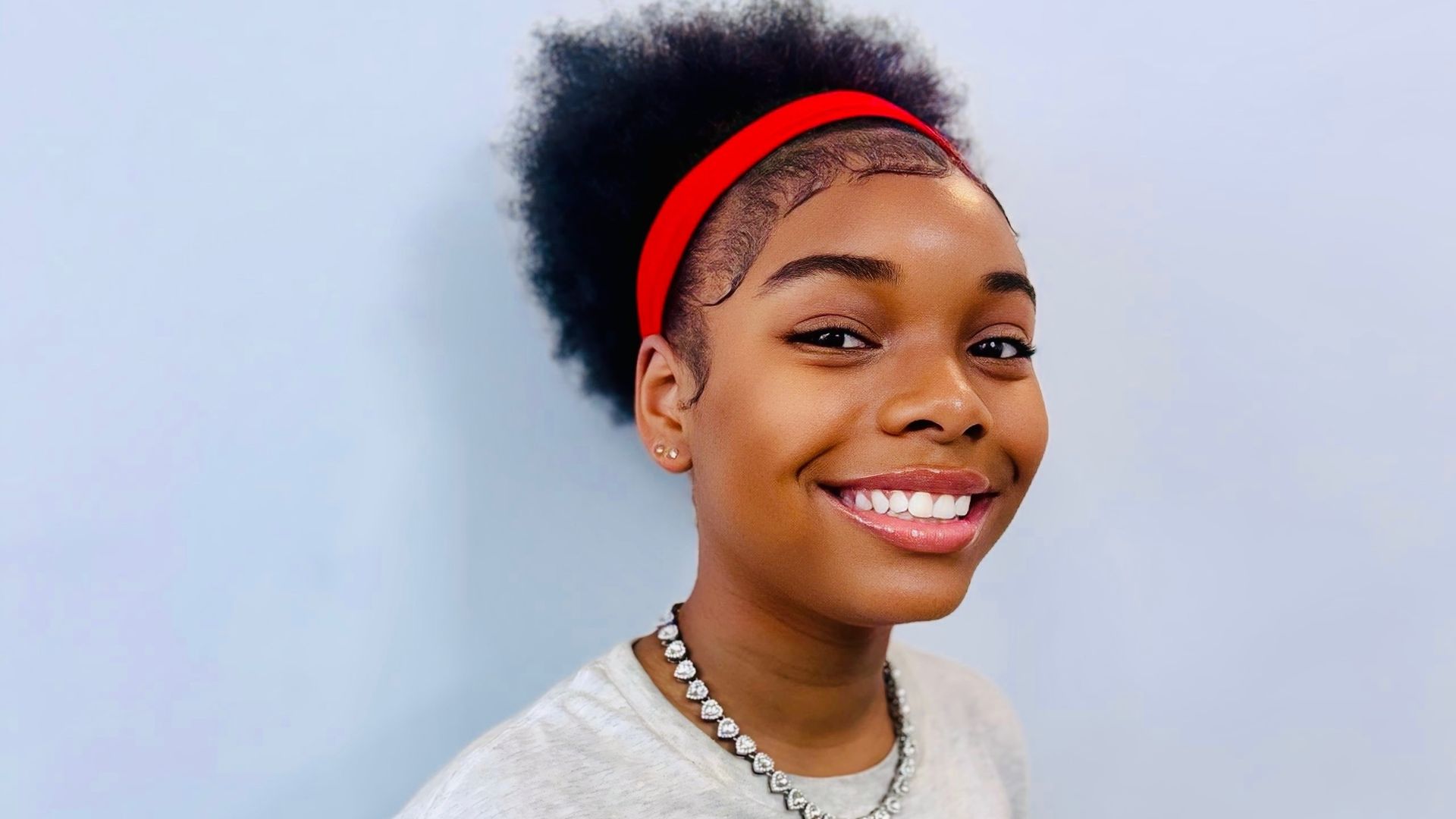 Portrait of a smiling young woman with natural hair pulled back by a red headband, wearing a silver necklace and a light gray top, against a pale blue background.