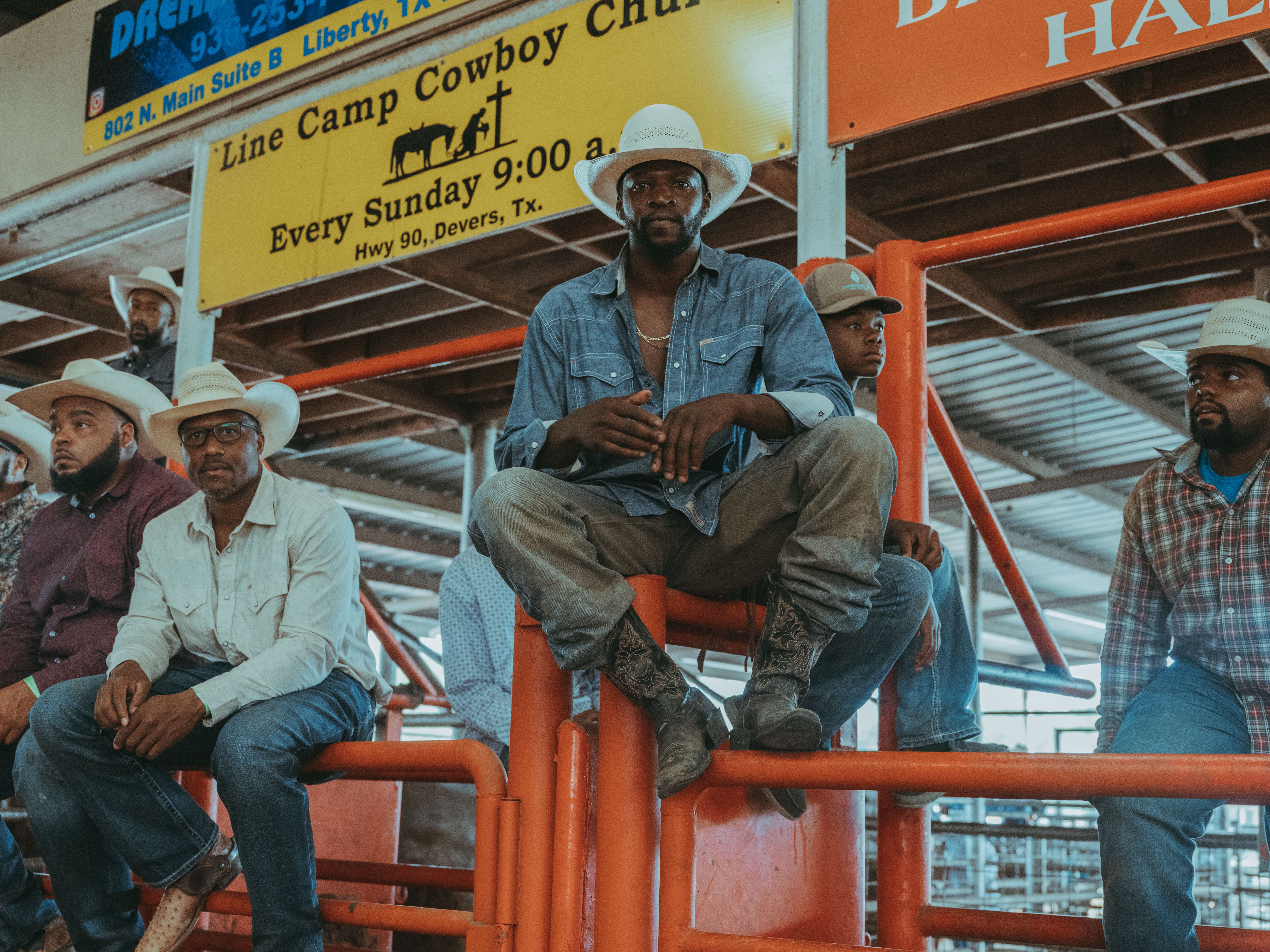 Photo of a group of Black men wearing cowboy hats and attire, sitting on a fence in a rodeo arena.