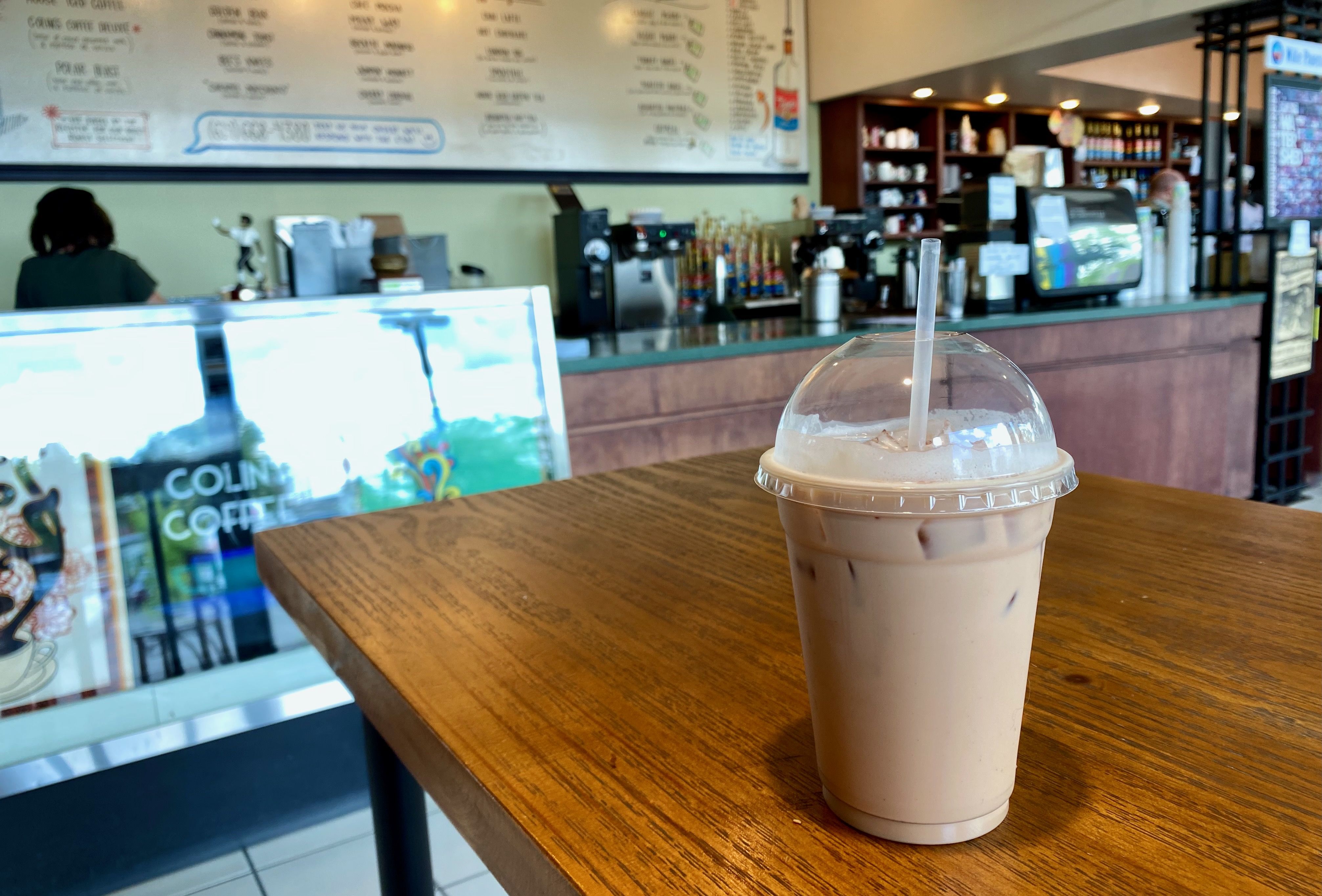 An iced mocha on a wooden table with a view of the barista counter in the background