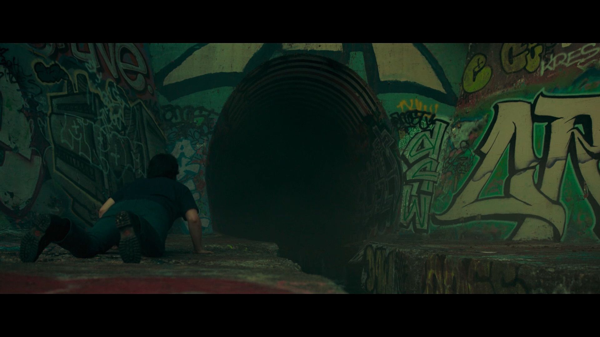 Person in a dark blue shirt crawls toward a large, dark tunnel mouth, surrounded by vibrant graffiti on green walls; a circular pipe spirals into the darkness.