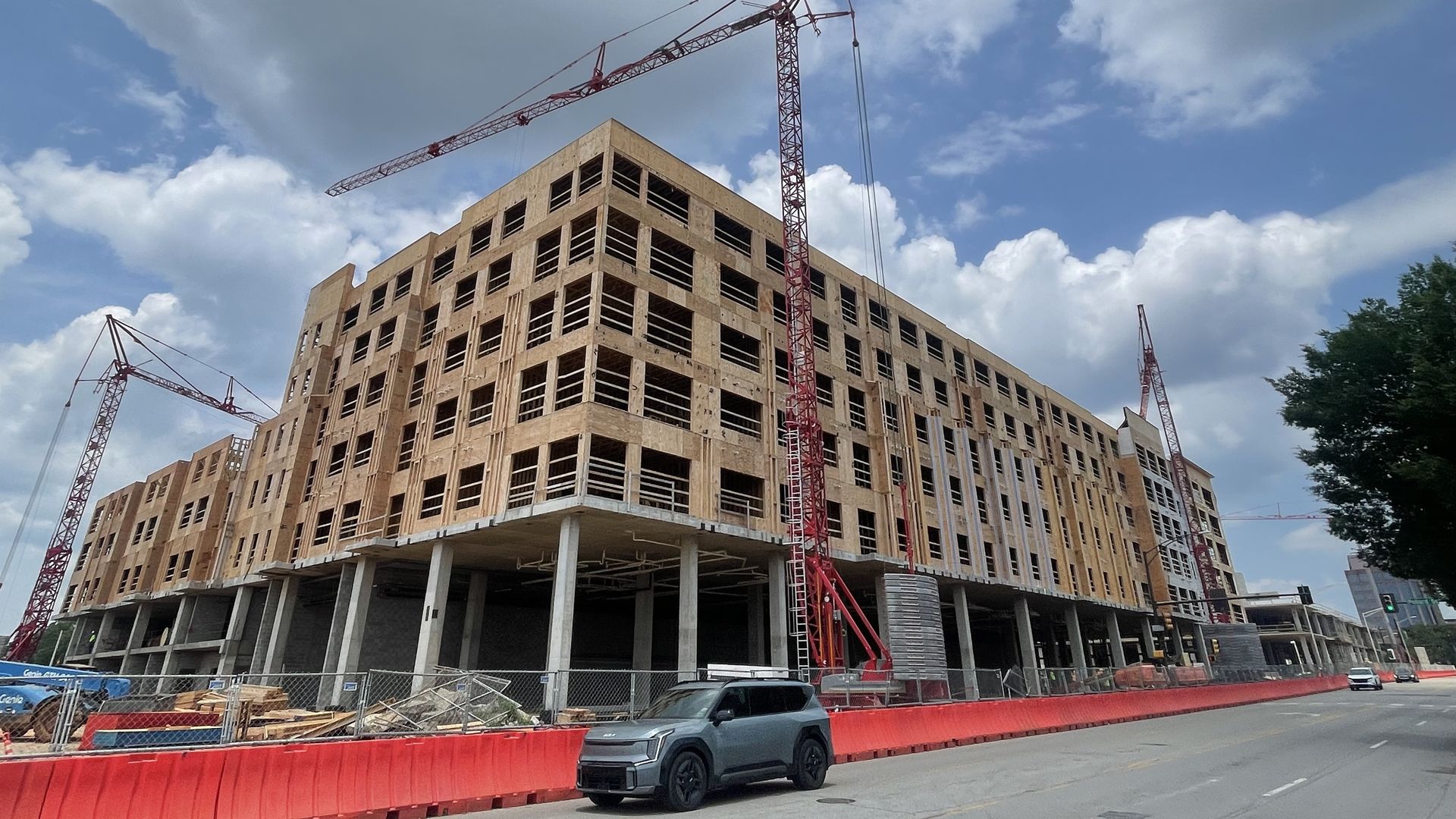 Red crane towers over a multi-story building under construction; wooden framing shows, blue sky with clouds, a red barrier along the street, and a gray SUV parked nearby.