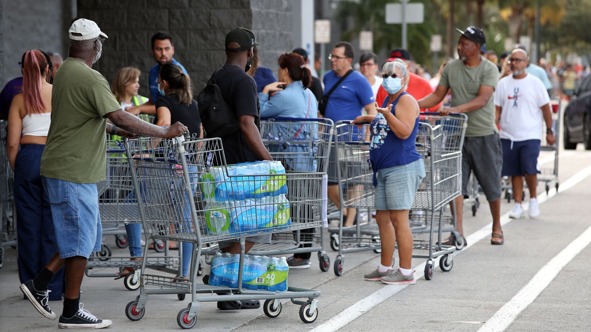 A long line of shoppers are seen past a person pushing cases of water outside a retail warehouse