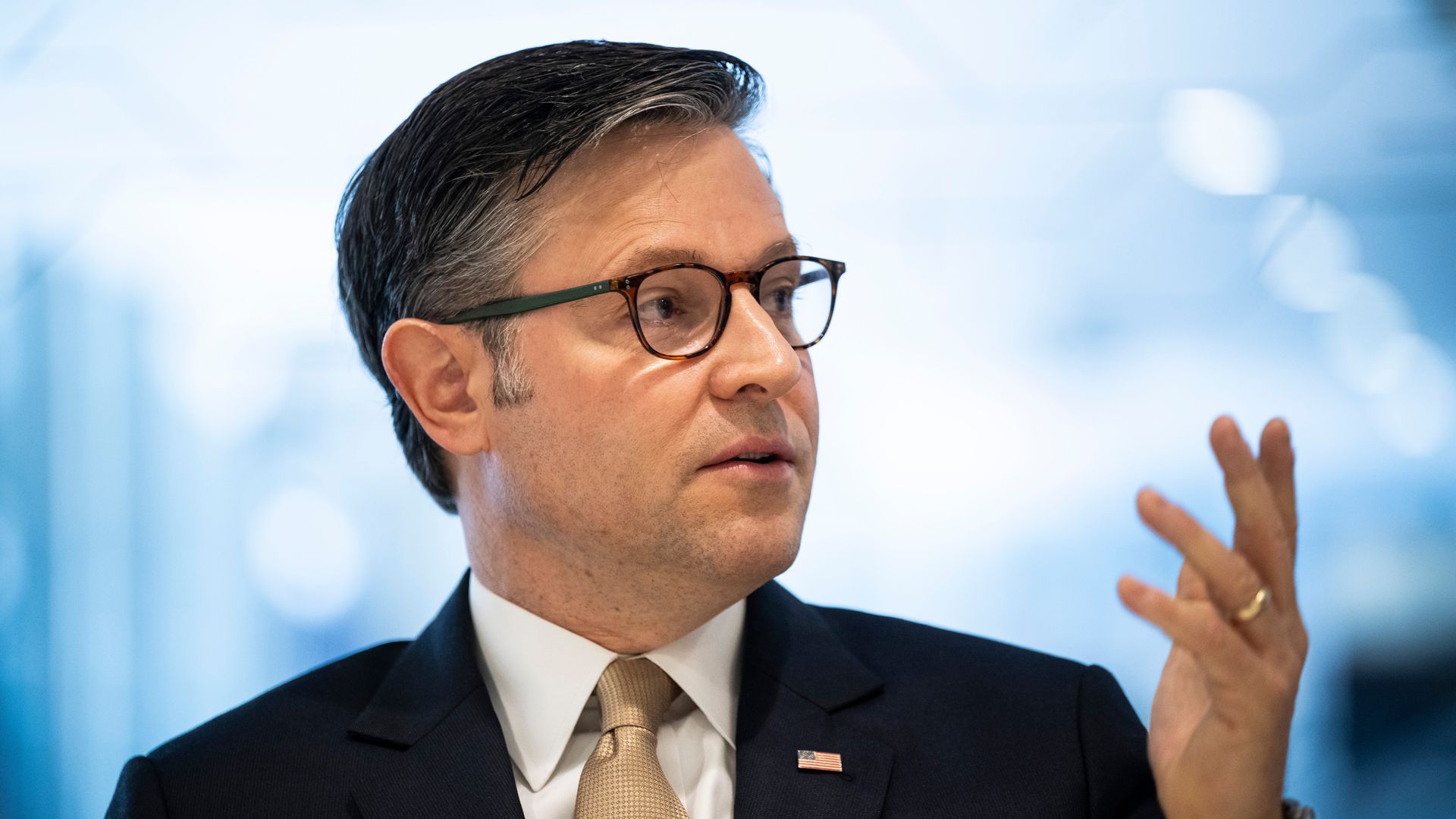 Man in a dark suit, beige tie, and glasses gestures with his right hand, wearing a U.S. flag pin on his lapel against a blurred blue-gray background.